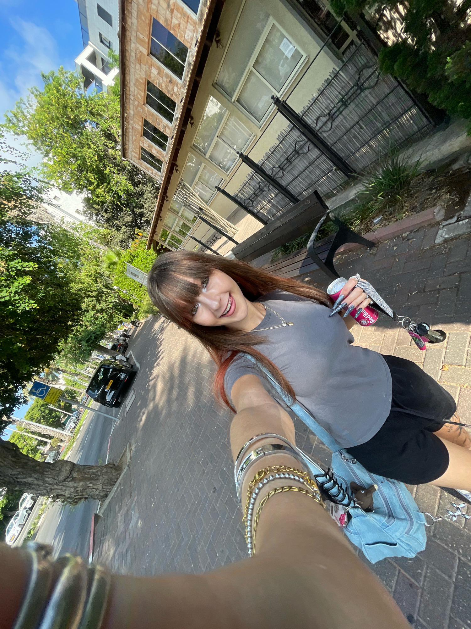 A young woman with long brown hair and bangs takes a selfie while standing on a brick sidewalk. She is wearing a grey t-shirt and black shorts. In her right hand, she holds a can of Red Bull and a set of keys. She is also wearing several bracelets on her left wrist. A light blue denim bag is slung over her shoulder.
Behind her, a building with beige stone siding and large rectangular windows stands. A black metal fence runs along the front of the building. In the background, trees line a street with parked cars. A blue sign indicating a disabled parking spot is visible on the left side of the street. The scene appears to be in the city of Raanana, Israel, on a sunny day.