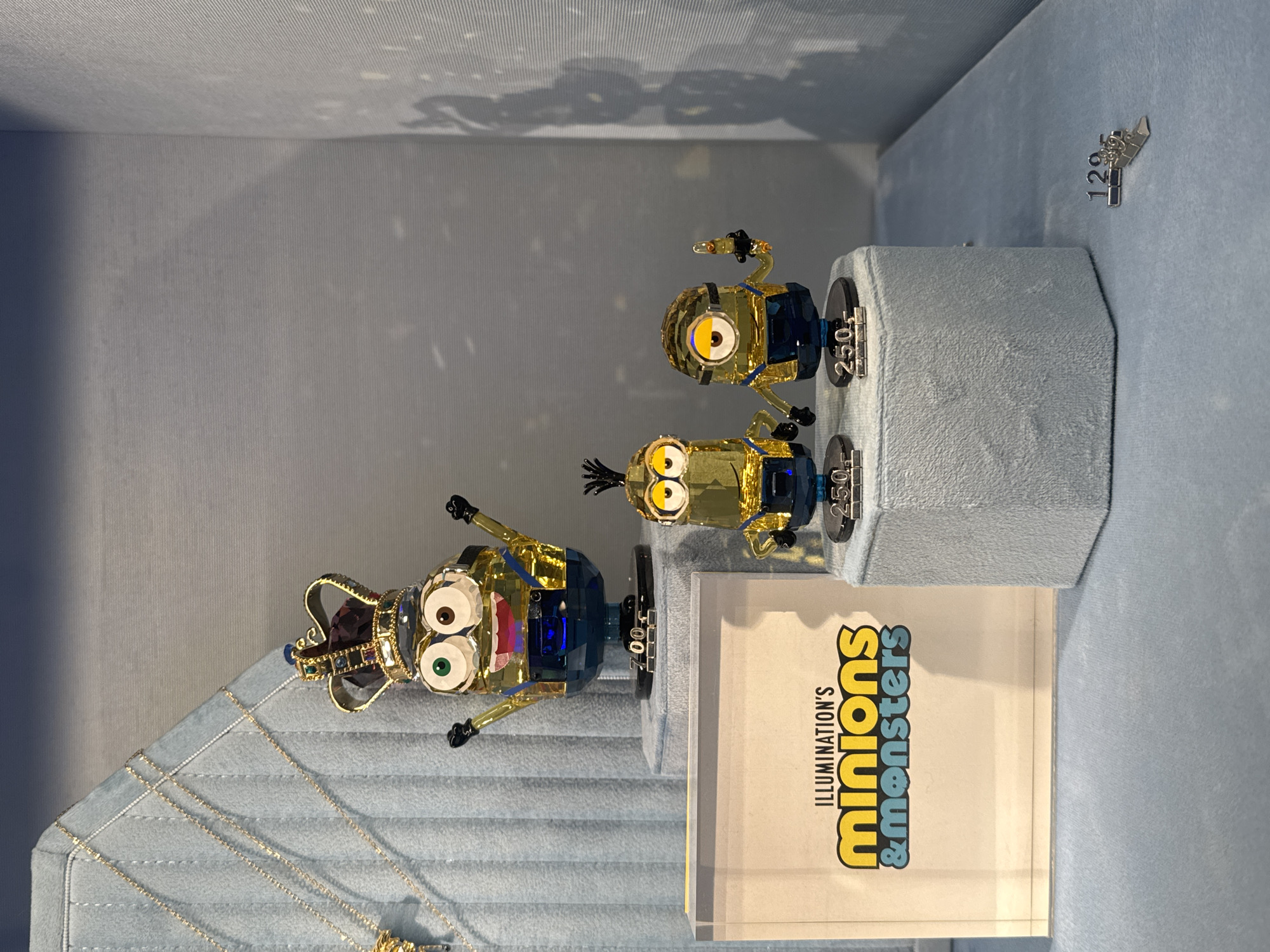 The image shows a retail display featuring crystal figurines of Minions characters from the "Illumination's Minions & Monsters" franchise. There are three figurines of different Minions characters on display, each with a price tag of 250.00. A fourth character is wearing a crown. The display also includes necklaces. The location is Spreitenbach, Switzerland.