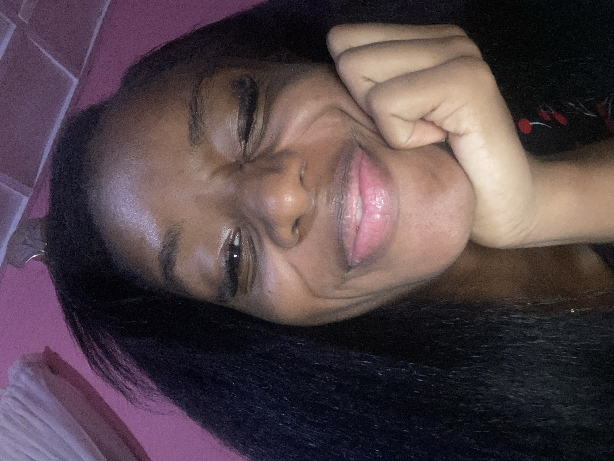 A close-up, selfie-style shot shows a young Black woman with long, dark hair. She is leaning her right cheek on her hand, with her fingers splayed against her chin and lower face. Her left eye is open, and her right eye is partially closed in a wink. She has light pink lip gloss on and appears to be smiling. The background is a pink wall, possibly textured, with a white towel or fabric draped in the upper left corner. The image is indoors and appears to be taken in a residential setting. There is no indication of the city of Jos, Nigeria, in the image.