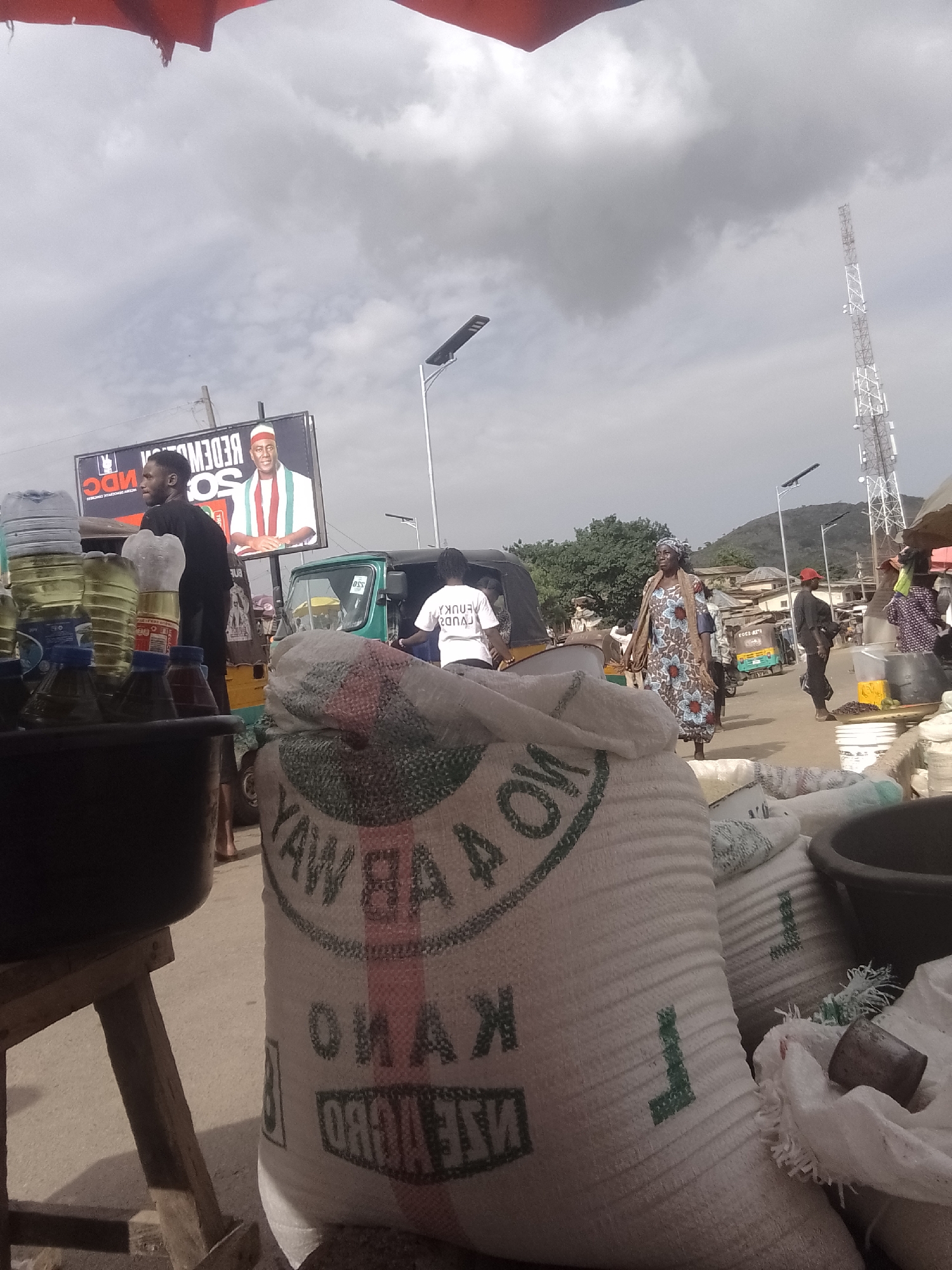 A medium shot shows a street market scene. In the foreground, a large white sack with green and red lettering is prominently displayed. To the left, a black bucket sits on a wooden stand. Behind these, a young man in a black t-shirt stands near a collection of plastic bottles filled with yellow liquid. Further in the background, a billboard features a man in traditional attire. A green motorized tricycle is parked on the street, and several people are visible walking and interacting. The environment appears to be a bustling urban market with a cloudy sky overhead and a distant cell tower.