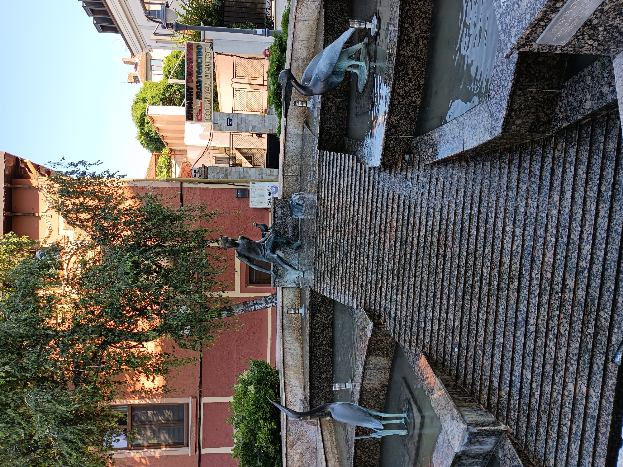 This image shows a tiered fountain with bronze sculptures of birds and a boy. The fountain is constructed of granite and has water flowing down its steps. In the background, there is a building with red and white facade and a sign in Cyrillic. This fountain is located in the city of Burgas, Bulgaria, specifically in front of the "Slovo" Bookstore, which is part of the "Koliren Center". The sculpture depicts a young boy reaching out to a bird, while two larger bird sculptures, a heron and a pelican, are positioned in separate pools. The scene is illuminated by sunlight, casting shadows on the fountain and the surrounding buildings.