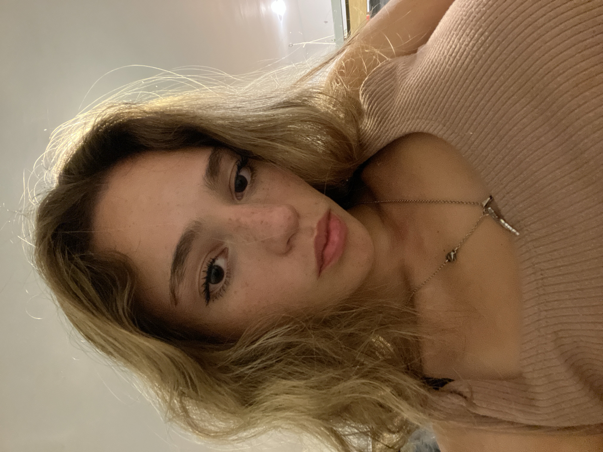 A young woman with blonde, wavy hair is looking at the camera. She has fair skin, freckles on her nose, and brown eyes. She is wearing a light pink ribbed shirt and a delicate silver necklace. The background is a blurry combination of white walls and ceiling. The photo appears to be taken indoors.