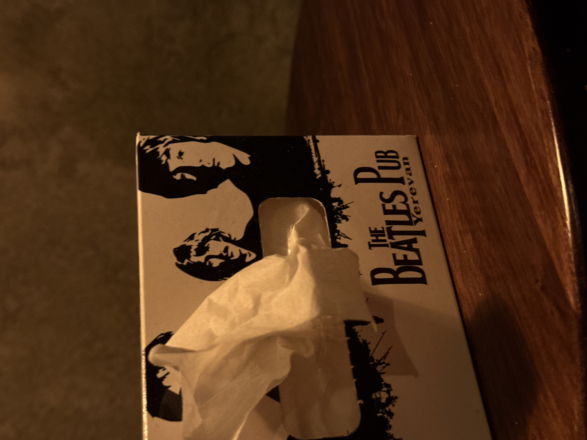 A tissue box with "THE BEATLES PUB Yerevan" printed on it sits on a wooden surface. The tissue box has a black and white graphic featuring the Beatles. A white tissue protrudes from the opening in the box. The background consists of a dark, blurry wall.