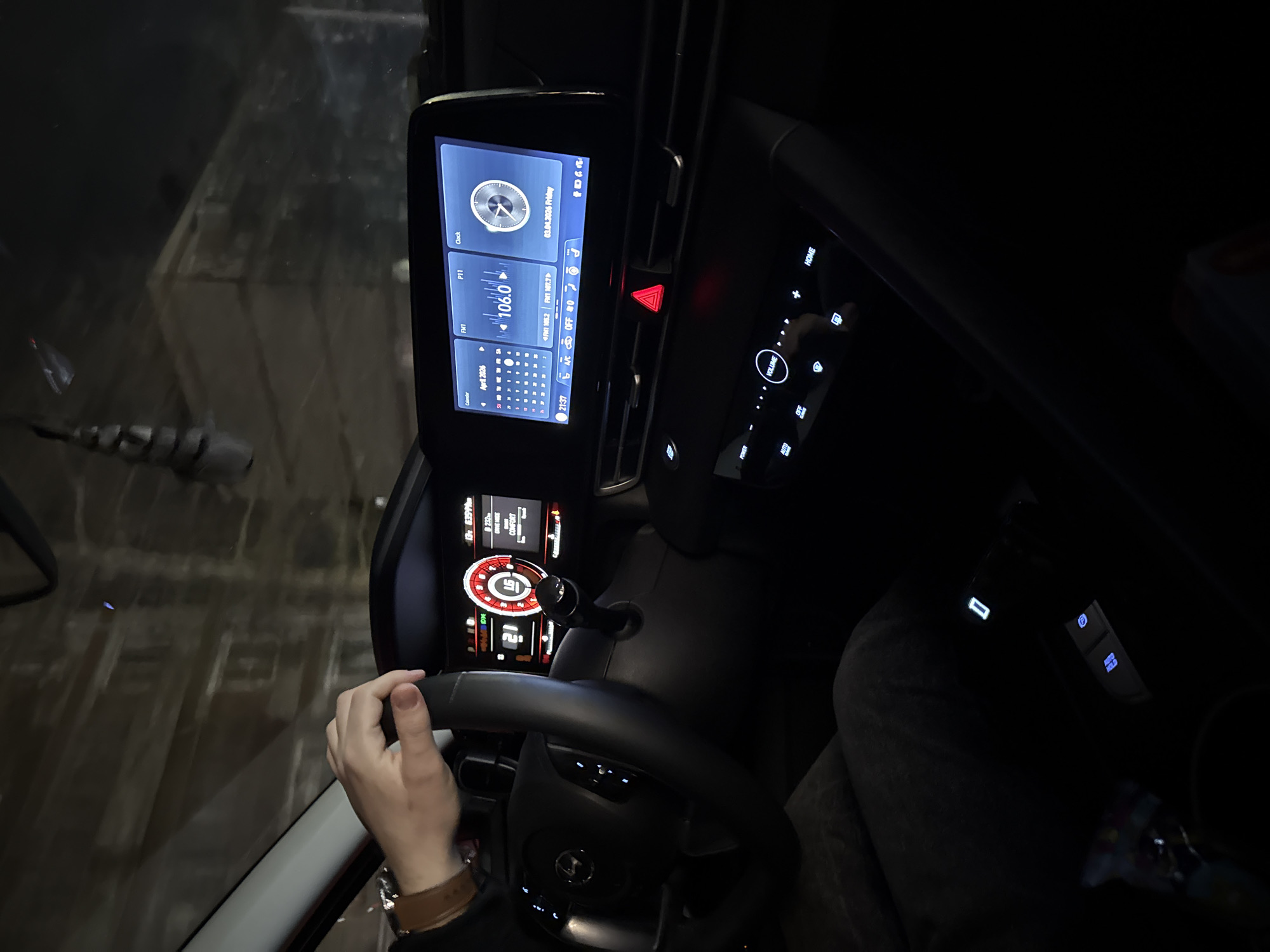 Here is a description of the image:
The photograph captures the interior of a vehicle at night. The dashboard is prominent, featuring an infotainment screen displaying various data, including a clock set to 21:37 on Friday, April 3, 2026, as well as radio information tuned to 106.0 FM. The climate control is set to AC off. To the right of the screen are illuminated buttons. The steering wheel is visible with a hand gripping it, suggesting the driver is in control. The instrument cluster behind the steering wheel shows additional information, including speed and RPM readings. The overall environment inside the car is dark, with the dashboard lights illuminating the immediate area. The car is driving in Unknown, Unknown, which is observable through the car's windshield.