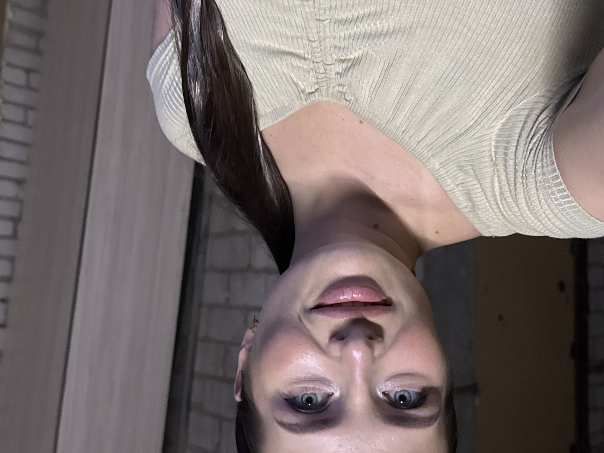 A woman is looking at the camera. She is wearing a light beige top. She has dark hair. The background includes a brick wall, a wooden wall, and a dark doorway. The location is in a city and the building could be an apartment.