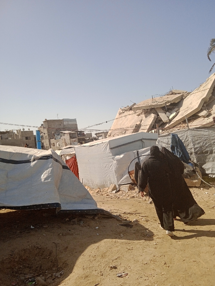 This image depicts an outdoor scene in Khan Yunis, Palestinian Territory, under a clear, pale blue sky. The ground in the foreground and midground is dry, sandy, and uneven, with scattered small debris.
In the mid-foreground on the right, two figures dressed in long, dark garments are walking away from the camera. The leading figure carries a large, circular metal object on her back, secured by a strap, and wears light-colored footwear.
The midground is occupied by several temporary shelters constructed from white tarpaulin material. One shelter on the left displays partial text "Gaza L" on its side. Another shelter shows a red fabric flap at its entrance. Piles of concrete rubble and assorted construction debris are present around these temporary structures.
The background features urban structures under varying conditions. On the right, a substantial pile of collapsed concrete building material, consistent with a multi-story structure, is visible, with exposed rebar. A fragment of a palm tree is discernible to the far right. To the left, a dense array of multi-story buildings extends into the distance, some appearing damaged. A faint, indistinct figure is visible in the distance near these buildings on the left.