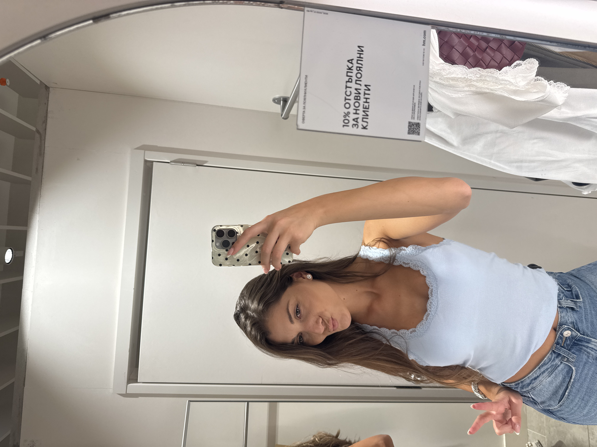 The image shows a young woman taking a selfie in a fitting room, likely at an H&M store in Burgas, Bulgaria. The woman has long brown hair and is wearing a light blue tank top and blue jeans. She is holding a smartphone with a speckled case to take the photo. The fitting room has a white door with a sign on it advertising a "10% discount for new loyal customers" in Bulgarian. There are clothes hanging in the background, including a white lacy item and a burgundy woven purse. The room appears to be clean and well-lit.