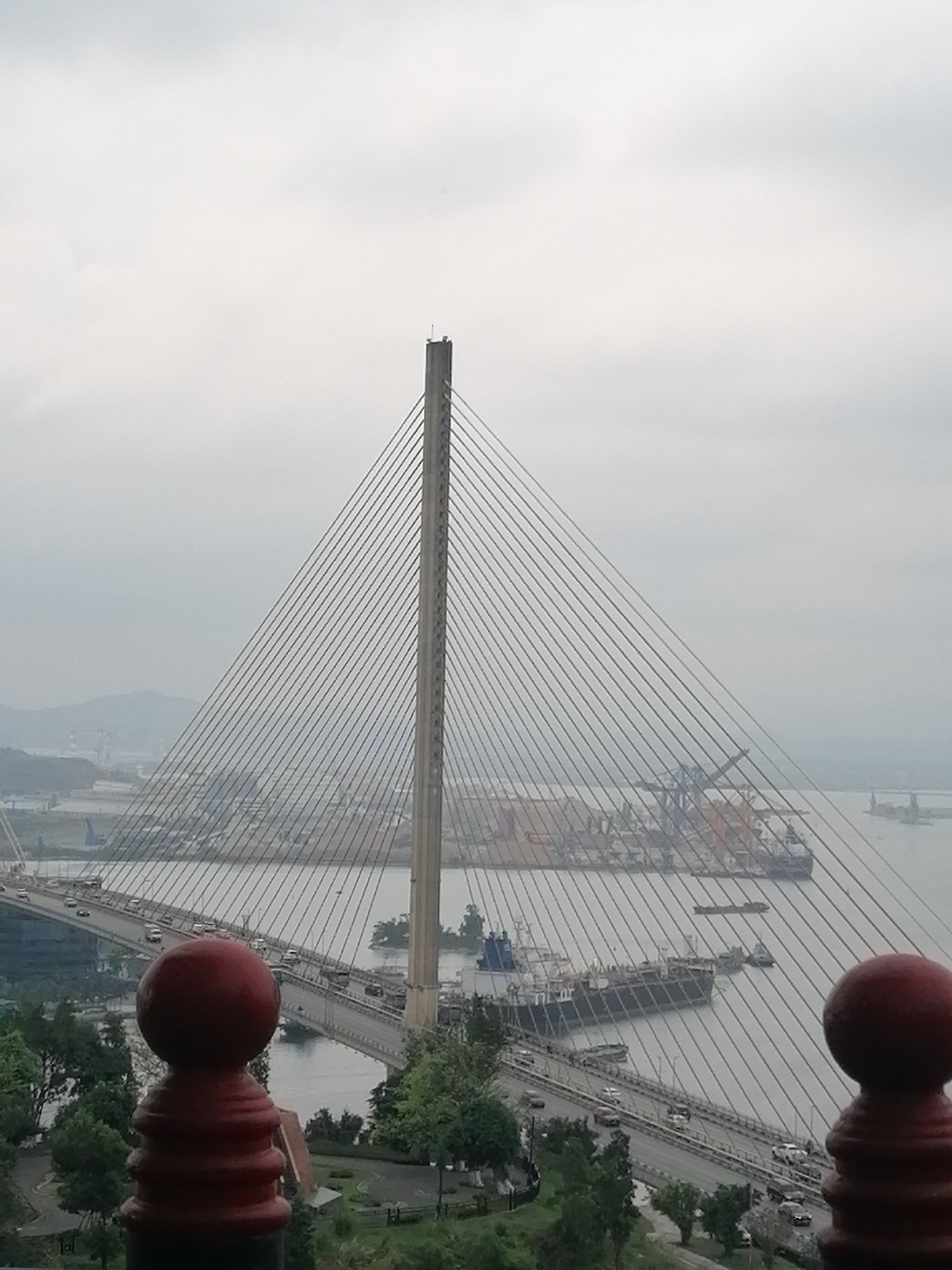 The image presents a view of a cable-stayed bridge in Hong Gai Ward, Vietnam. The bridge is seen through a foreground featuring a reddish-brown, decorative railing. The bridge spans a body of water where ships and boats are visible. A port area with industrial cranes and large cargo ships can be seen beyond the bridge. Traffic is moving on the bridge. The sky is overcast, creating a muted color palette. The foreground also includes greenery, suggesting a park or landscaped area.