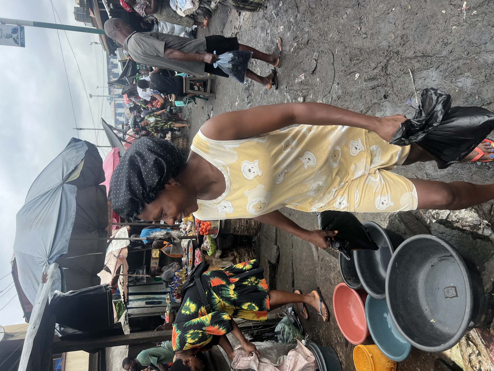 In Nkpor, Nigeria, a bustling marketplace teems with activity under a cloudy sky. A woman in a black polka-dot headwrap and a yellow sleeveless top with a cartoon bear print, along with matching yellow shorts, is captured mid-stride, holding a black plastic bag. She is walking towards the right of the frame, her expression lively and engaged, perhaps in conversation with someone out of view.
To her left, another woman in a vibrant, multi-colored floral dress is bent over, reaching into a large grey plastic basin filled with a white substance, possibly rice or flour, and holding a similar black plastic bag. The ground is a muddy, uneven surface, indicating recent rainfall or poor drainage, scattered with various plastic basins of different sizes and colors, including yellow, red, teal, and dark grey.
In the background, a chaotic yet organized market scene unfolds. Stalls draped with fabrics and tarpaulins offer a variety of goods, including vegetables and bottles of liquid. Several other individuals are visible, some tending to their stalls, others browsing or walking through the crowded aisles. The general atmosphere is one of commerce and daily life in a busy Nigerian urban setting.