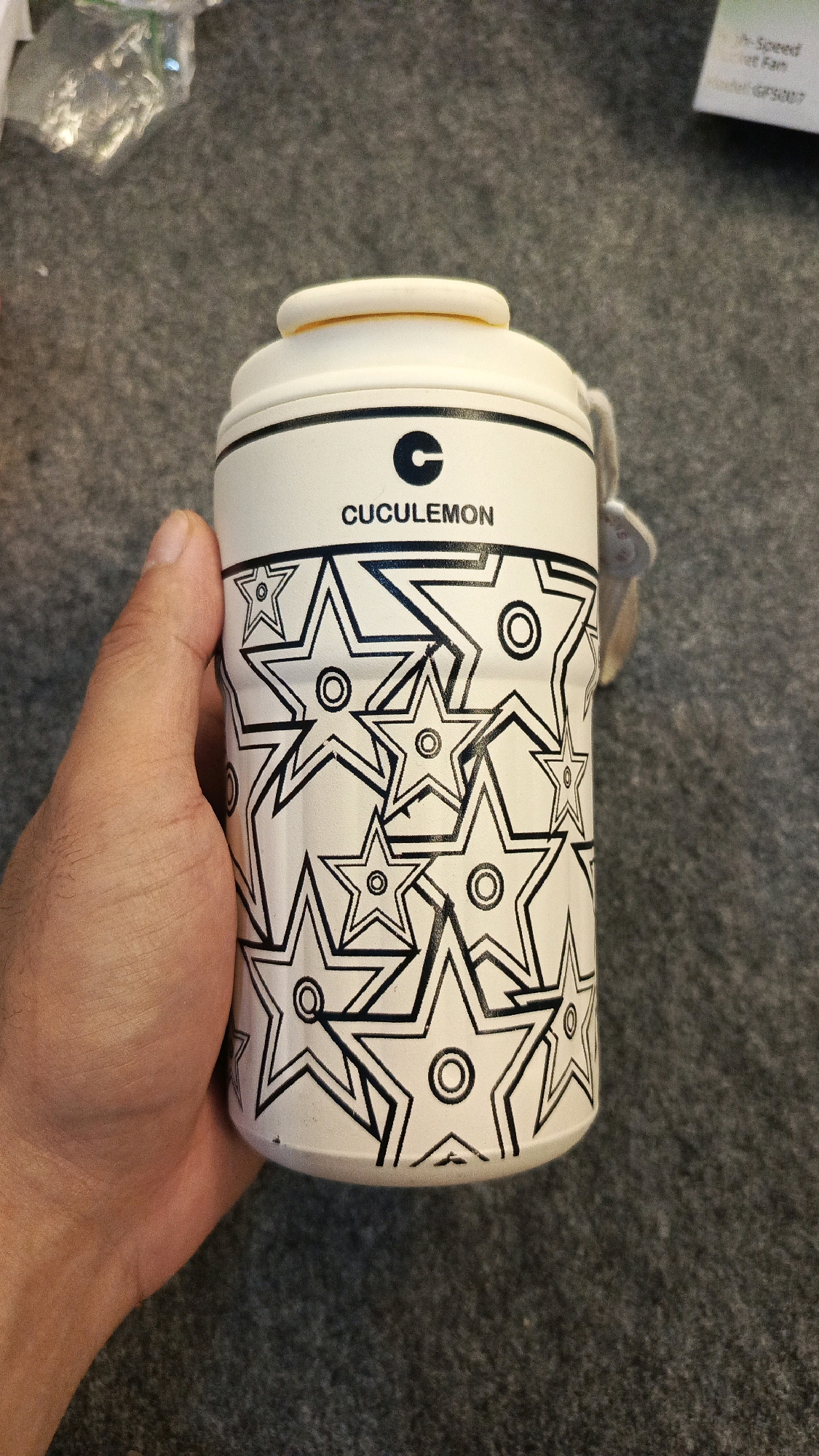 A hand is holding a white cylindrical object, possibly a travel mug or a thermos, decorated with black star patterns. The object has a white lid with a yellow ring. Above the stars, the word "CUCULEMON" is printed in black letters, preceded by a black stylized "C" logo. The background is a textured grey surface, likely a carpet or fabric. A partially visible label with some text in English is in the upper right corner. The image is well-lit, with the object in focus. There is no discernible connection to Babadan, Indonesia.