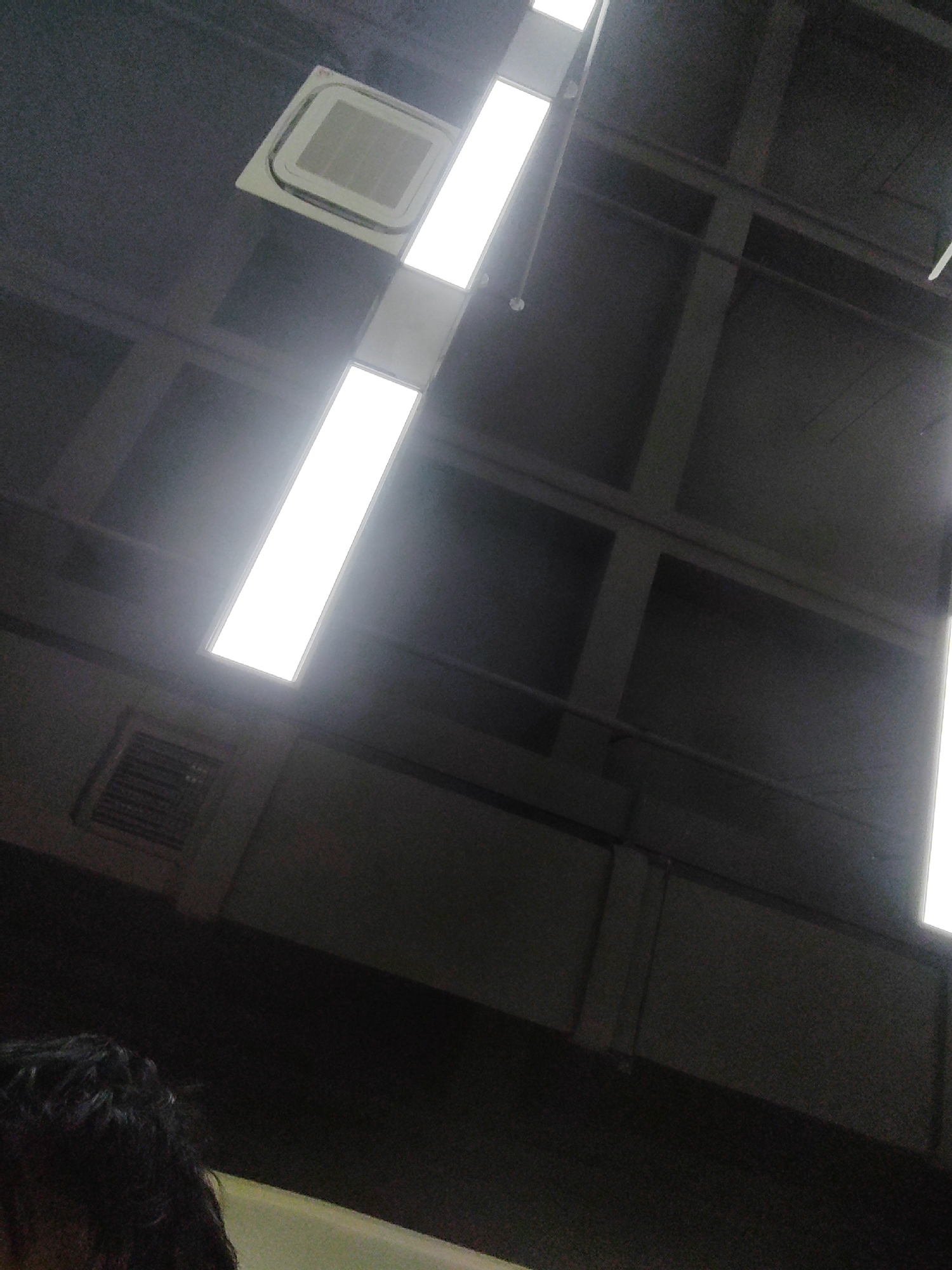 The image shows an indoor scene, likely a hallway or large room with a textured ceiling. A prominent rectangular light fixture with a bright white glow is visible, casting light downwards. To the left of the light fixture, a square ventilation grille is embedded in the ceiling. The ceiling structure appears to be made of dark beams and panels. In the lower left corner, a portion of a person's dark, curly hair and shoulder is visible. The overall lighting is dim, with the bright light fixture being the primary source of illumination in the frame. The location of Chongqing High-tech Zone, China, is not depicted in this image.