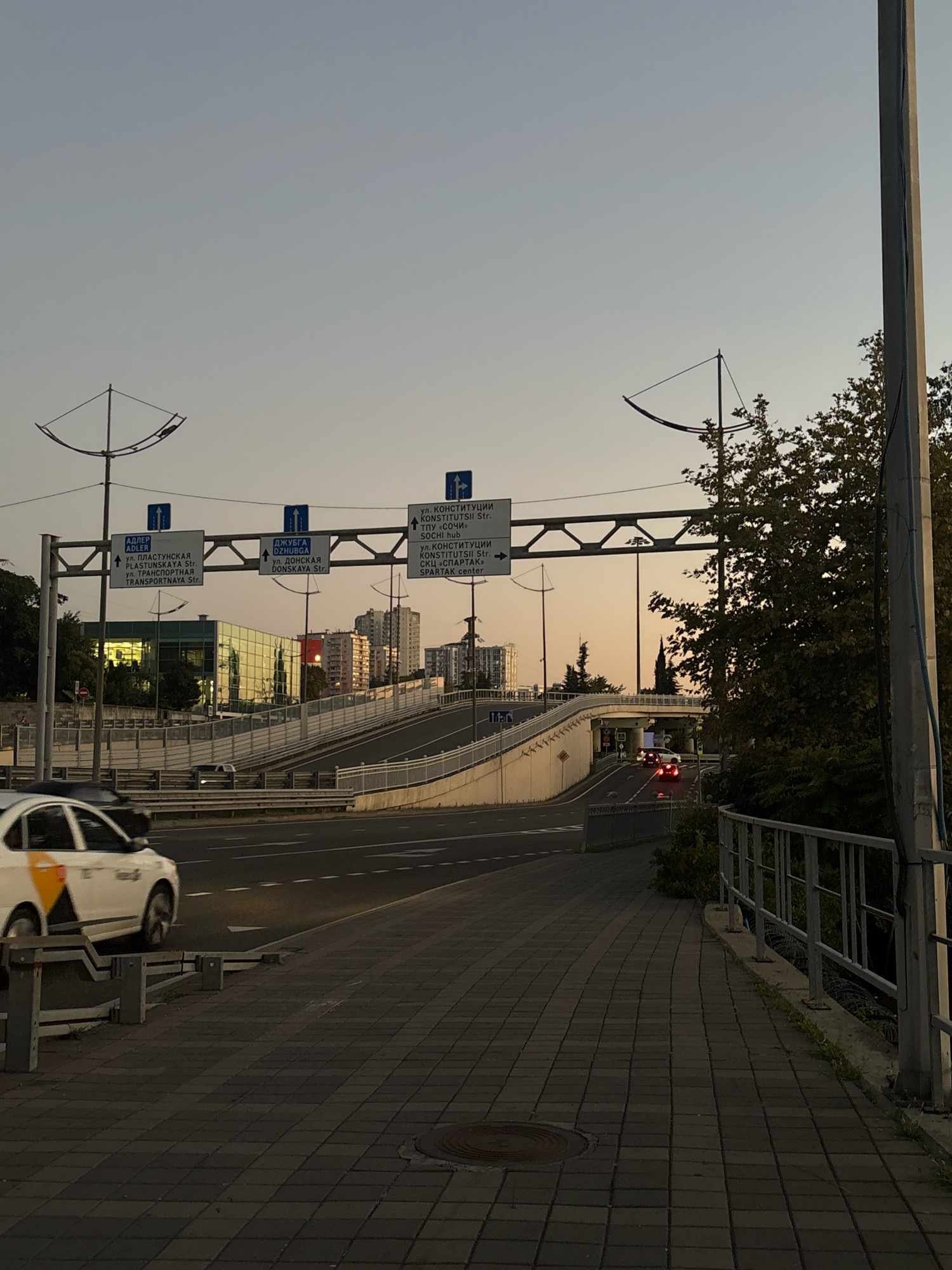 The image shows an urban scene in Sochi, Russia, likely at dusk. A multi-level road system is visible, with cars on the road. There are directional signs in Russian indicating directions to Adler, Dzhubga, and Constitution Street in Sochi, as well as "Sochi hub" and "Spartak center". A modern building with a glass facade is situated on the left side of the image, reflecting the sky. A pedestrian walkway paved with dark gray tiles runs along the right side of the scene, bordered by a metal railing. Trees and greenery are present in the background, and the sky is a pale orange and blue, indicating the transition from day to night. A white car with yellow and black markings is in motion on the road in the foreground.