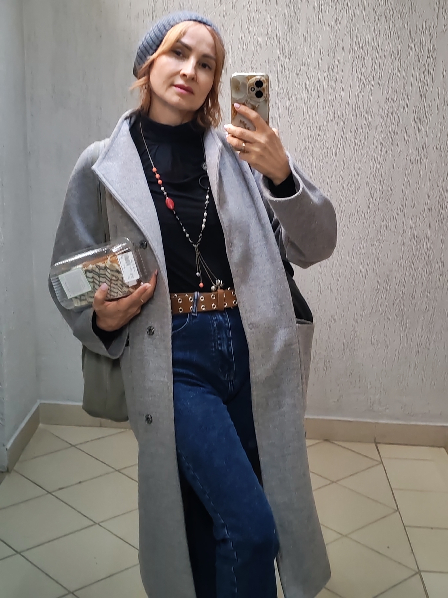 A woman takes a mirror selfie indoors, holding a smartphone in her right hand and a clear plastic container of pastries in her left. She has light brown hair, visible beneath a grey knit beanie. Her expression is neutral as she looks towards the camera.
Her attire includes a black turtleneck, a long grey overcoat/cardigan, and dark blue jeans. A distinctive brown belt with multiple metal studs or eyelets cinches her waist. She wears layered necklaces, one featuring red/coral-colored beads and another with white, pearl-like beads and metallic accents. Her fingernails are painted a light pink.
The background consists of a light-colored, textured wall, resembling stucco or painted concrete. The floor is covered with light-toned square tiles arranged diagonally, separated by darker grout lines. A beige baseboard runs along the bottom of the wall, and a corner is visible to the left, suggesting a hallway or common area within a building, likely in Chaikovsky, Russia. The clear plastic container holds several pastries or rolls and has white labels affixed to it, though the text, including what might be "ШТ" (pcs/pieces), is largely illegible. The smartphone has a gold/beige patterned case with a prominent dual-lens camera system.