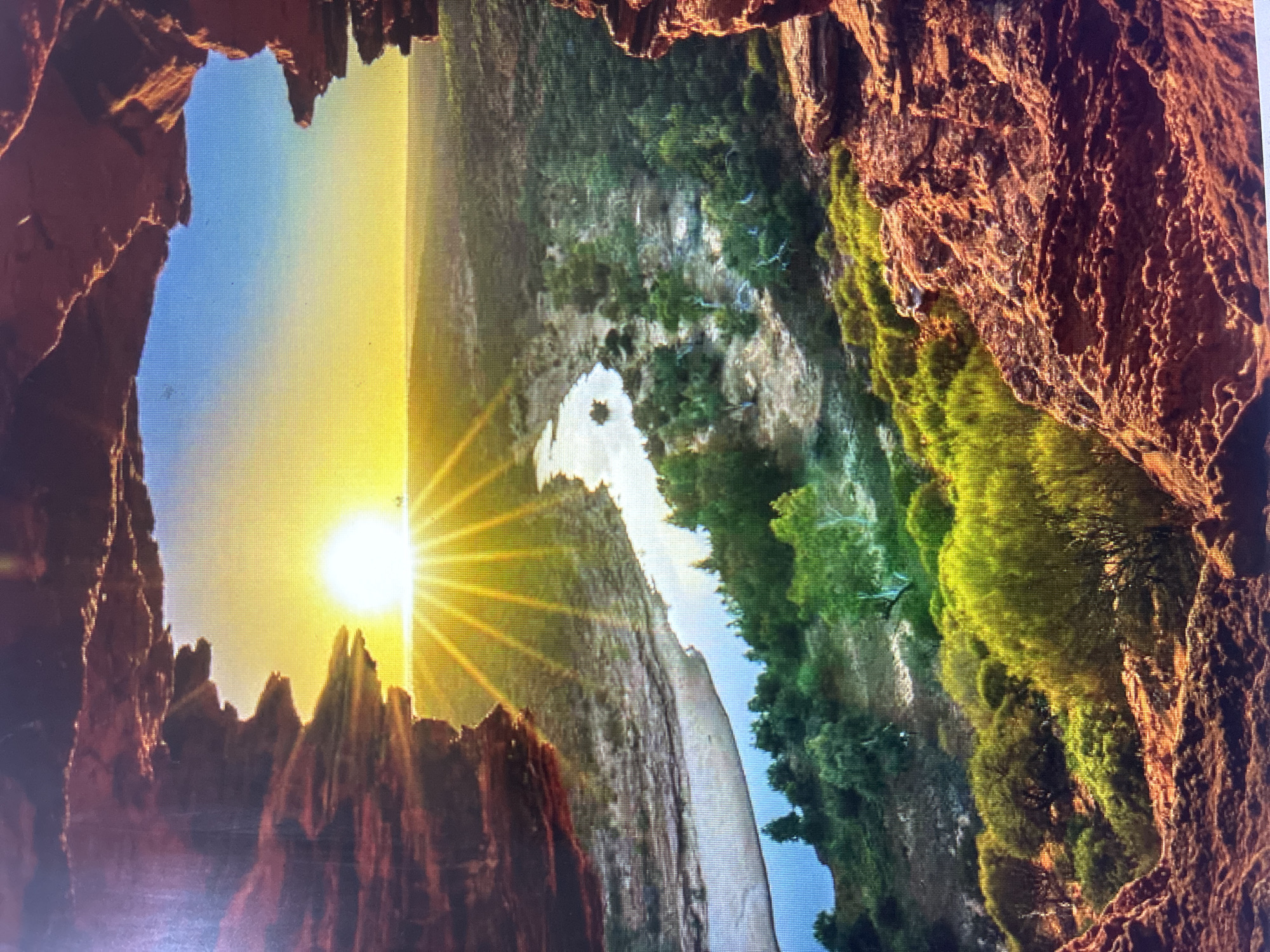 The image displays a dramatic natural landscape, captured at either sunrise or sunset, viewed from within a large, rugged rock formation or cave entrance. The scene is framed by dark reddish-brown, heavily textured rocks that form a jagged opening on the left, top, and right sides of the image.
A brilliant, low-lying sun is positioned on the left, casting intense golden rays that radiate across the sky. The sky transitions smoothly from a bright yellow-orange near the sun to a clear, soft blue further away.
Through the rock aperture, a verdant valley or canyon unfolds. A winding river or stream meanders through the middle ground, its banks rich with lush green vegetation and trees that also cover the steep slopes and cliffs on the right side of the valley. A distinct white, possibly sandy or rocky, formation is visible within or alongside the river.
There are no people or visible text in the image. The strong sunlight, contrasting with the shadowed rock frame, creates a sense of depth and awe. The rugged rock formations and the river valley with dense foliage are characteristic of mountainous regions, consistent with landscapes found in and around Asenovgrad, Bulgaria.