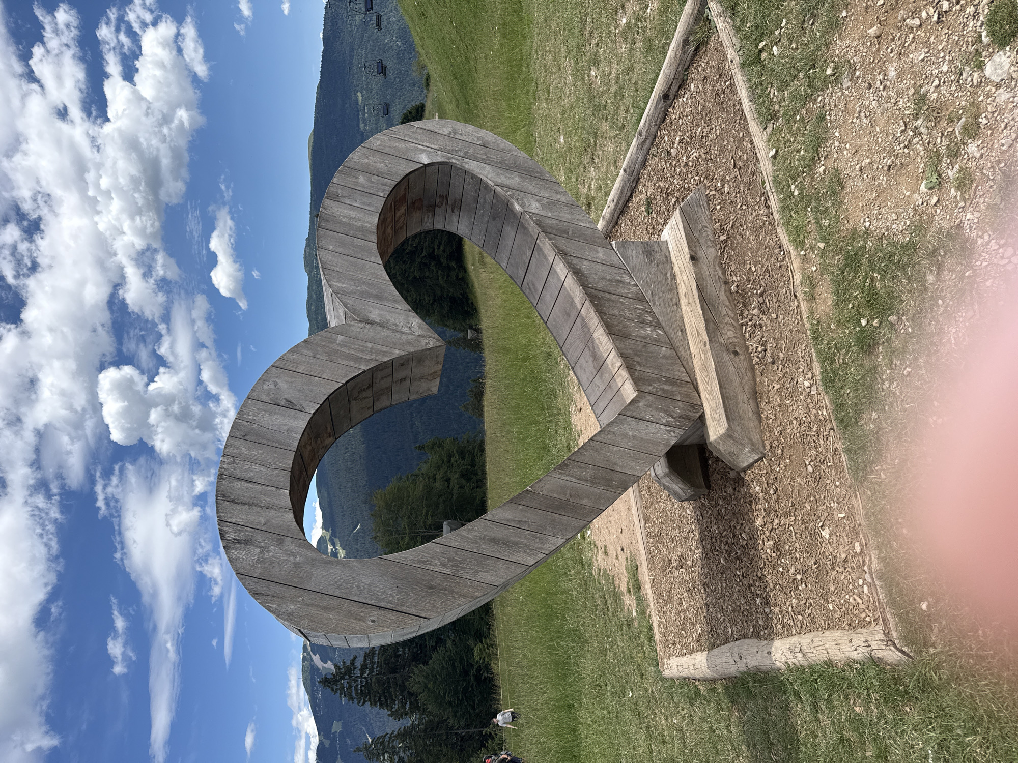 An outdoor, eye-level, medium shot captures a large, weathered wooden heart sculpture positioned in a grassy area at the foot of a mountain. The sculpture, made of planks, is supported by wooden beams and rests on a bed of wood chips bordered by logs. In the background, lush green grass covers rolling hills, leading to a dense forest of pine trees. Beyond the trees, the rugged slopes of a mountain range are visible, with a ski lift extending up one side. The sky is a vibrant blue, dotted with fluffy white clouds. Two distant figures are seen walking on the grass to the left of the sculpture. The scene is well-lit by natural daylight.
