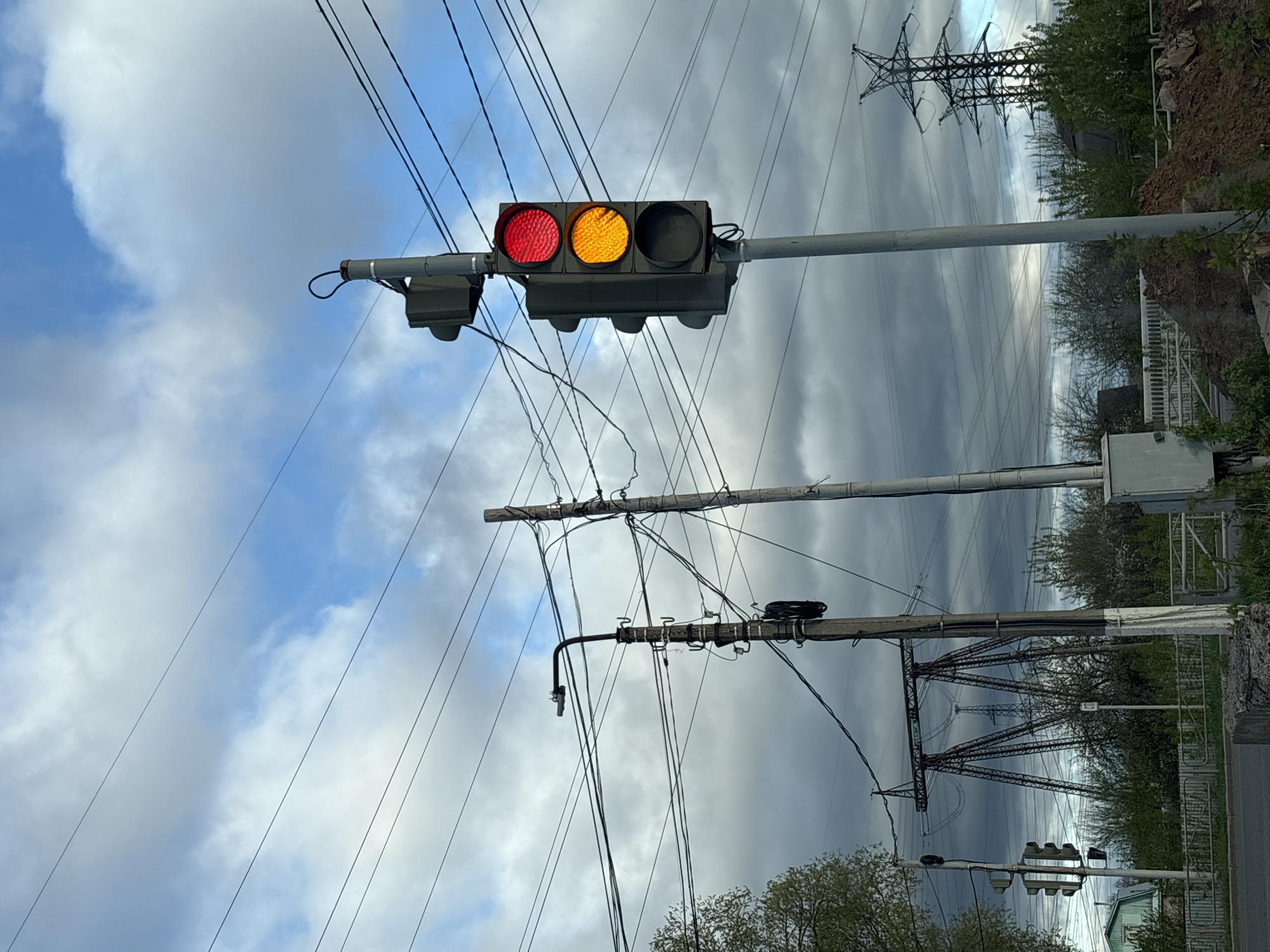 The image shows a traffic light in Chaikovsky, Russia. The red and yellow lights are illuminated. The traffic light is mounted on a gray pole. Overhead power lines and a transmission tower are visible in the background. The sky is cloudy with patches of blue. Vegetation is present along the right side of the image.
