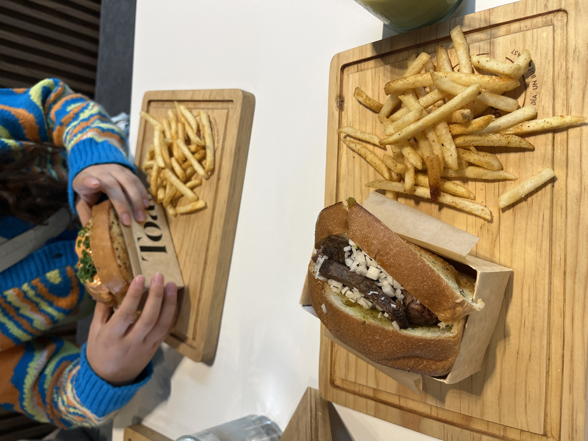 In San Bernardino Tlaxcalancingo, Mexico, on a white table, two wooden trays hold servings of food. One tray in the foreground contains a hot dog-like sandwich filled with meat and diced onions, accompanied by a pile of french fries. The sandwich is presented in a small cardboard holder lined with parchment paper. The second tray, positioned further back and slightly out of focus, holds another sandwich, partially visible, topped with a creamy sauce and green garnish, also served in a cardboard holder, and a portion of french fries. A person wearing a brightly colored sweater with blue, orange, and yellow stripes is visible in the background, holding the sandwich on the second tray. To the left of the foreground tray, a glass of water is partially visible. The lighting is natural, suggesting an indoor setting during the daytime.
