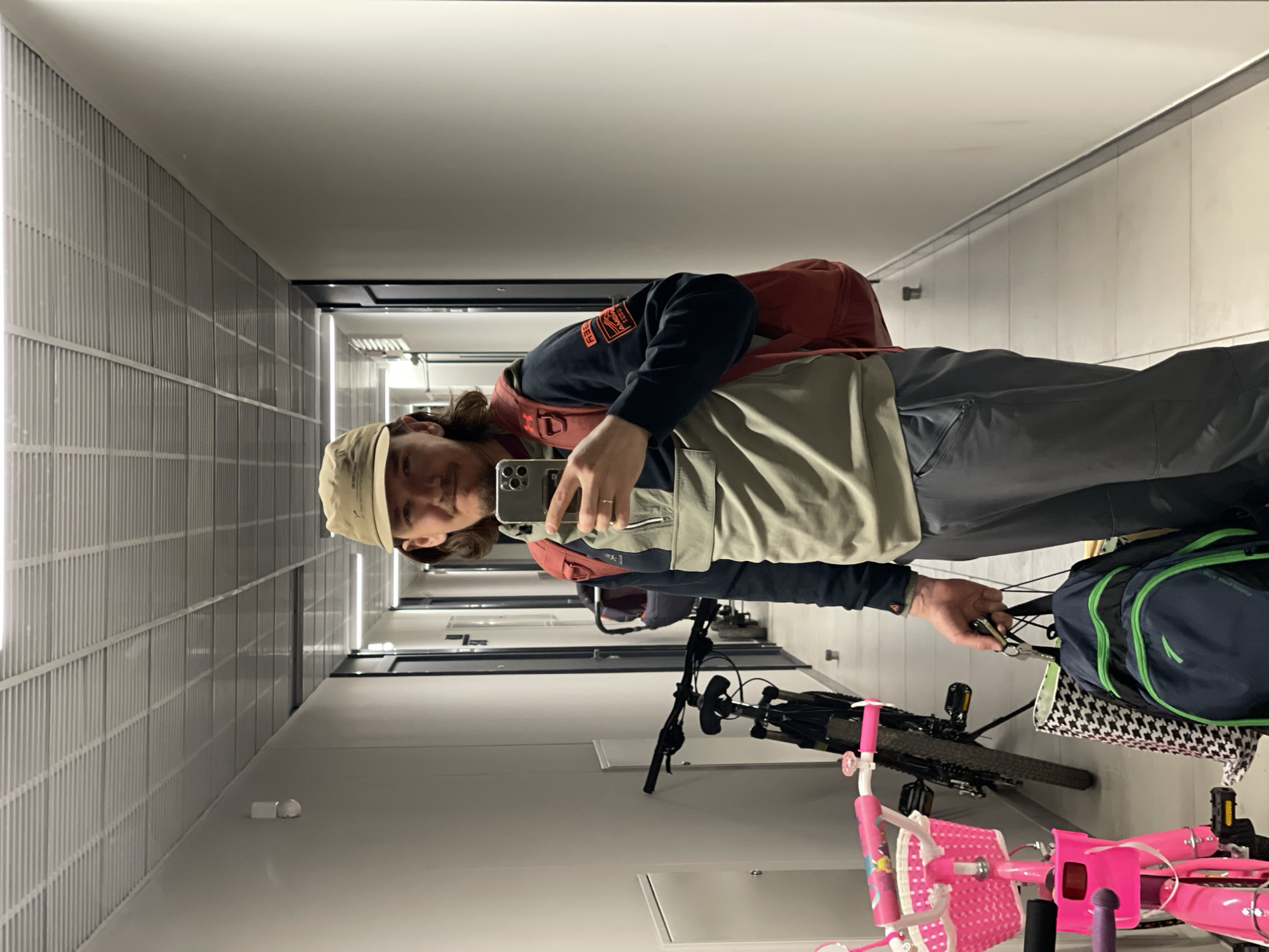 A person is taking a selfie in a long, narrow hallway with a textured wall on the left. The person wears a tan baseball cap, a navy blue long-sleeved shirt with orange lettering, a tan jacket, and grey pants. They are holding a smartphone in their right hand, presumably to take the picture. A red backpack is visible on their back. A black and grey mountain bike is standing next to a pink child's bicycle. There is a green and blue backpack on the mountain bike. The hallway has doors on the right and extends into the distance. The location is likely an apartment building, but specific information about Izhevsk, Russia, is not available.