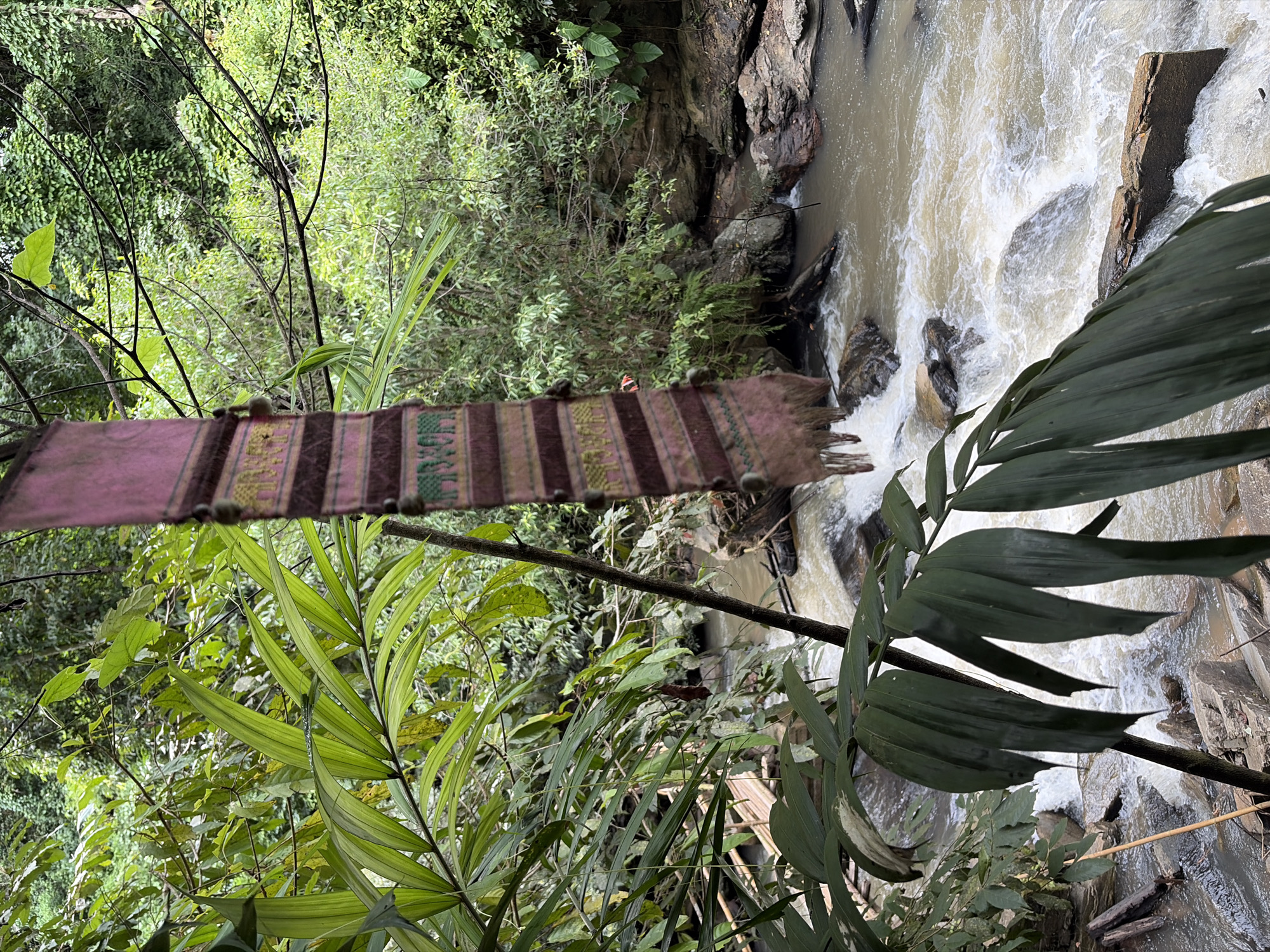 In Ban Huai Takhrai, Thailand, a narrow, woven textile hangs suspended in front of a natural scene. The textile, predominantly pink with alternating horizontal bands of brown and green, features an intricate pattern of elephants woven into the green stripes. The bottom edge of the textile frays into tassels. Behind this hanging object, lush green foliage dominates the background. Various types of leaves, from broad palm-like fronds in the foreground to smaller, dense leaves further back, create a vibrant canopy. A natural stream flows rapidly over rocks in the midground. The water is turbulent, with white foam indicating its swift movement. Rocks of various sizes are visible along the stream bed and banks, some covered in moss. The environment suggests a tropical, possibly jungle-like setting. The lighting is diffused, consistent with an overcast day or dense forest cover. There are no people visible in the image.