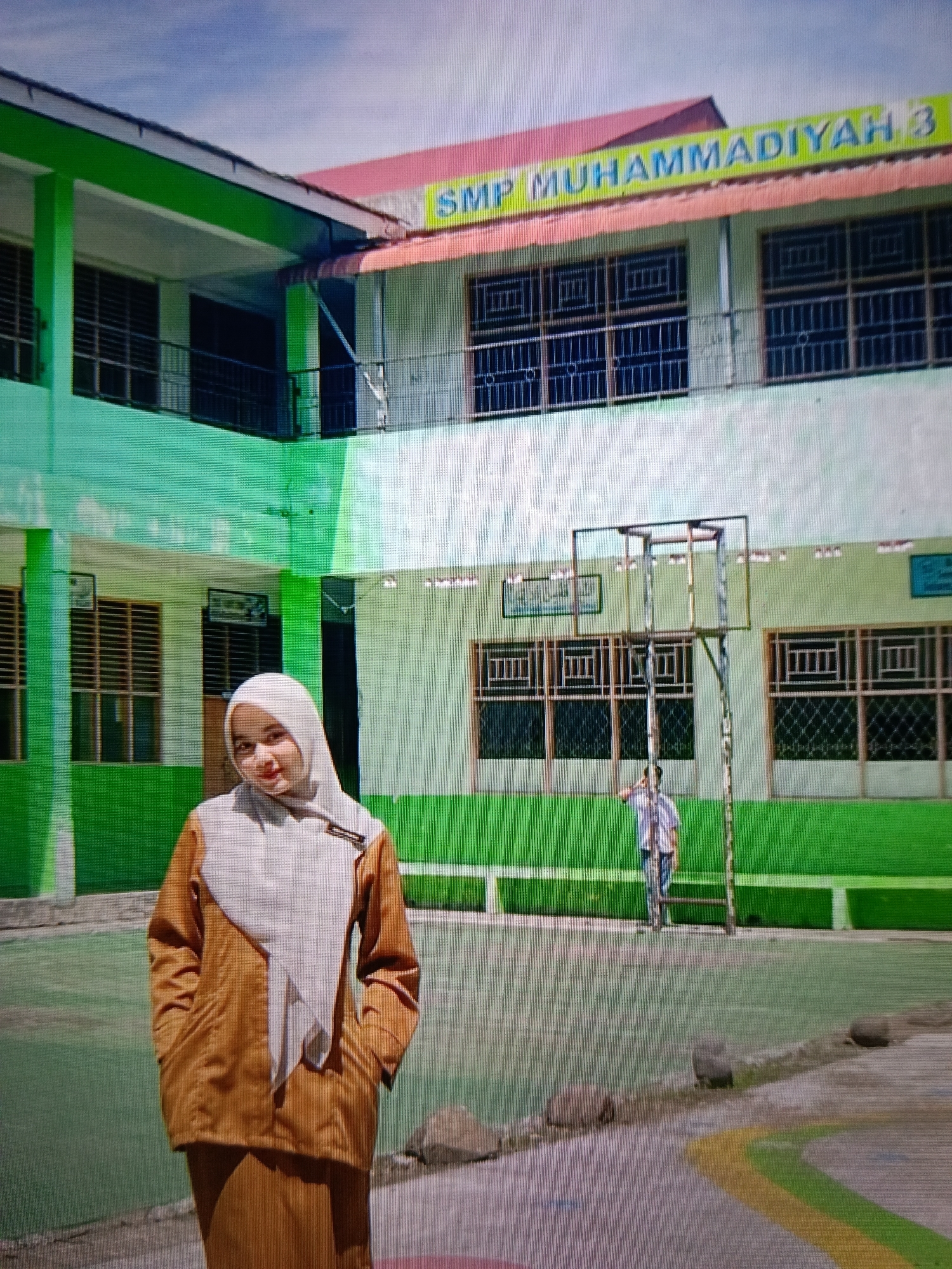 A young woman wearing a light brown hijab and an orange outfit stands in the foreground, facing the camera with her hands in her pockets. She is in front of a building with a red roof and green trim. The building has multiple windows and a sign that reads "SMP MUHAMMADIYAH 3". In the background, a basketball hoop is visible, and a person in a striped shirt and jeans is standing near it. The scene appears to be a schoolyard in Raha, Indonesia.
