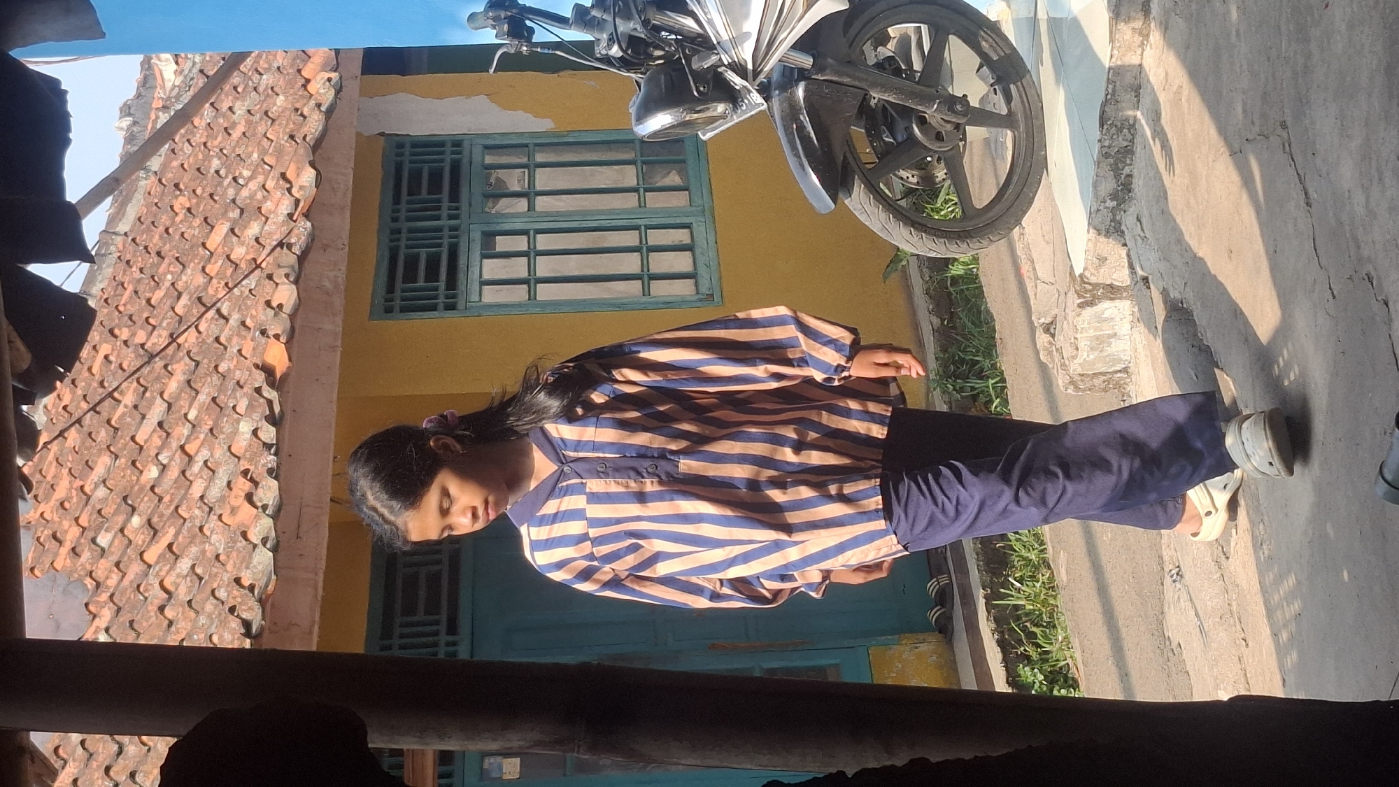 A young girl with dark hair pulled back is standing and looking down. She is wearing a long-sleeved striped shirt in shades of blue and tan, with a collared neckline and buttons down the front. She is also wearing dark blue pants. She has on light-colored slip-on shoes. The girl is standing on a concrete path in front of a yellow building with a tiled roof. A green-framed window is visible on the building. To the right of the girl, a motorcycle with a black seat and silver accents is parked. The ground is made of cracked concrete and there is some sparse green vegetation. The scene appears to be outdoors in daylight, with strong shadows indicating bright sunlight. The location is identified as Rahong, Indonesia.
