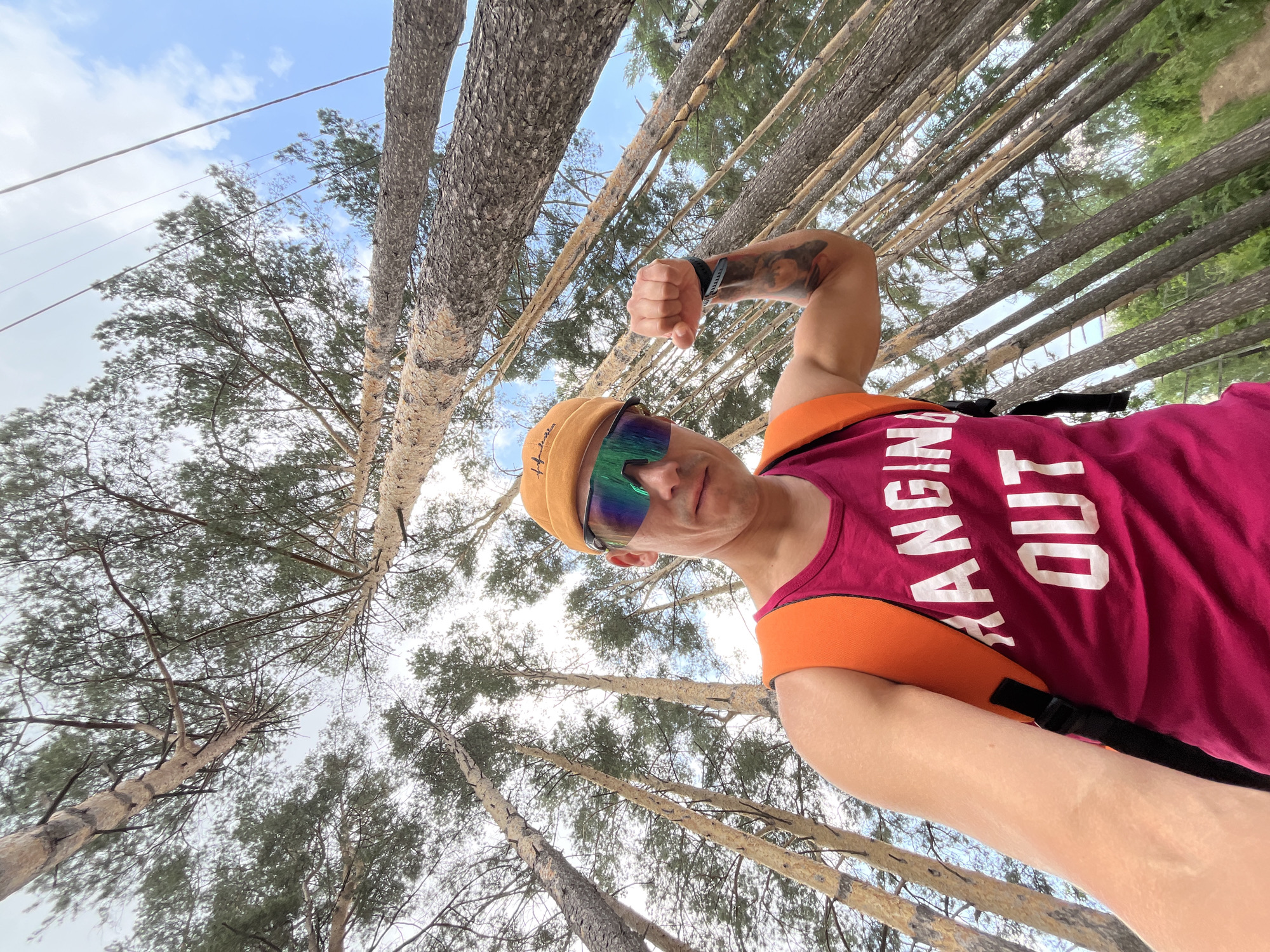 In the city of Chaikovsky, Russia, a low-angle, wide shot captures a man in a bright orange beanie and mirrored sunglasses, flexing his bicep. He is wearing a magenta tank top with white text that reads "HANGIN OUT" and an orange backpack. The man is positioned in the foreground, with his body angled towards the camera. Behind him, a dense forest of tall, slender pine trees stretches upwards towards a bright blue sky with scattered white clouds. The trees are closely packed, creating a sense of depth and immersion in the natural environment. The ground beneath the trees is not clearly visible, but hints of green foliage suggest a forest floor. The lighting indicates it is daytime, with sunlight filtering through the canopy, casting subtle shadows. The perspective of the shot emphasizes the height of the trees and the vastness of the forest.