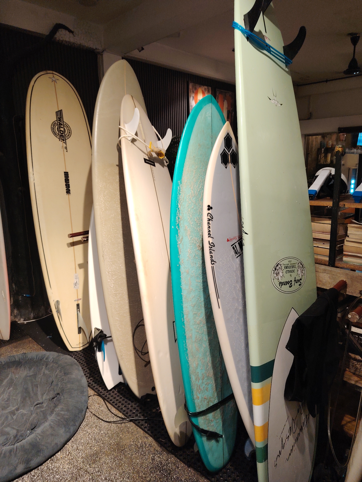 An indoor scene displays a collection of approximately nine surfboards, arranged vertically and leaning against a dark background wall and a rack structure. The surfboards exhibit various colors, lengths, and conditions, indicative of different types and usage.
On the left, a cream-colored longboard prominently features a black "WALDEN SURFBOARDS" logo. To its right, a white surfboard is fitted with two white fins, secured by white plastic fin holders and a yellow strap. Further right, a heavily waxed light blue/teal surfboard has a black leash attached to its tail. Behind it, a light grey surfboard bears "Channel Islands" branding and a black hexagonal fin system logo.
A prominent pale green surfboard on the right side displays black fins secured by a blue strap at its upper section. This board also features an oval "DONALD TAKAYAMA HAWAIIAN DESIGNS SURF BOARDS USA" sticker and horizontal stripes (dark green, yellow, white, light green) on its lower half. A partially visible white surfboard on the far right shows handwritten text "Hawaiian Designs." Many boards show signs of use, including wax residue and minor scuffs.
The floor in the foreground is light grey concrete, transitioning to a dark, perforated matting beneath the surfboards. A grey, circular fabric cushion or pet bed rests on the concrete floor in the lower-left corner. Black electrical cords are visible on the floor near the boards. In the background, stacks of light-colored rectangular objects, possibly books or boxes, are visible on shelves to the right. A dark ceiling fan hangs from an off-white ceiling in the upper-right background. The background wall on the left is dark and appears to have vertical corrugations. The lighting is artificial, creating shadows. The environment suggests an indoor retail space, such as a surf shop, or a storage area. The specific location is in Unknown, Unknown.