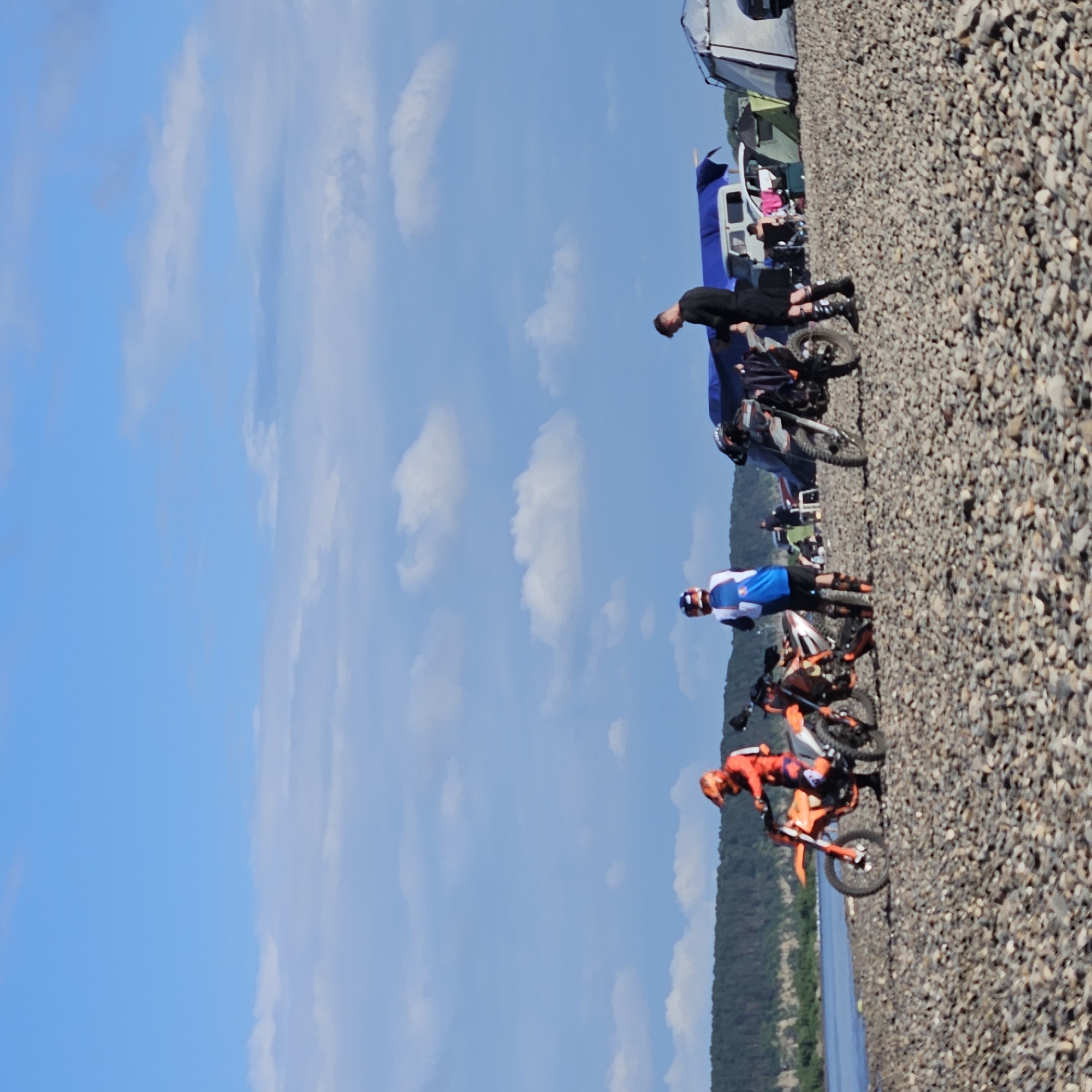A group of people are gathered on a rocky shore next to a body of water, with a forested hill in the background. The sky is blue with scattered clouds. Several dirt bikes are parked on the shore, and some of the people are wearing riding gear. Tents and vehicles are also visible in the background, suggesting a campsite or gathering. The scene is consistent with an outdoor recreational activity, possibly involving off-road motorcycling. The location is identified as the city of Elykaevo, Russia.