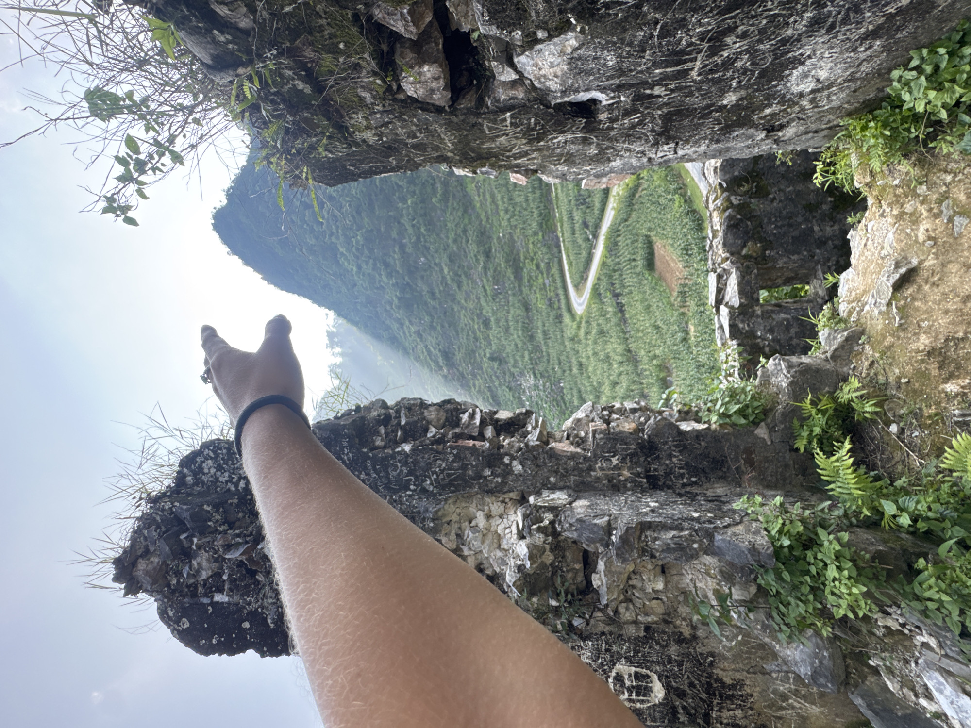A person's arm extends from the lower left corner into a scene depicting a view from a ruined stone structure. The arm, clad in a dark wristband and a ring, is positioned against the backdrop of a mountainous landscape. The ruined structure frames the view, with weathered stones and vegetation lining the edges of the opening. Beyond the stones, a road winds through a lush green valley, ascending towards a mountain in the distance. The scene suggests the location to be in the Đường Thượng Commune, Vietnam.