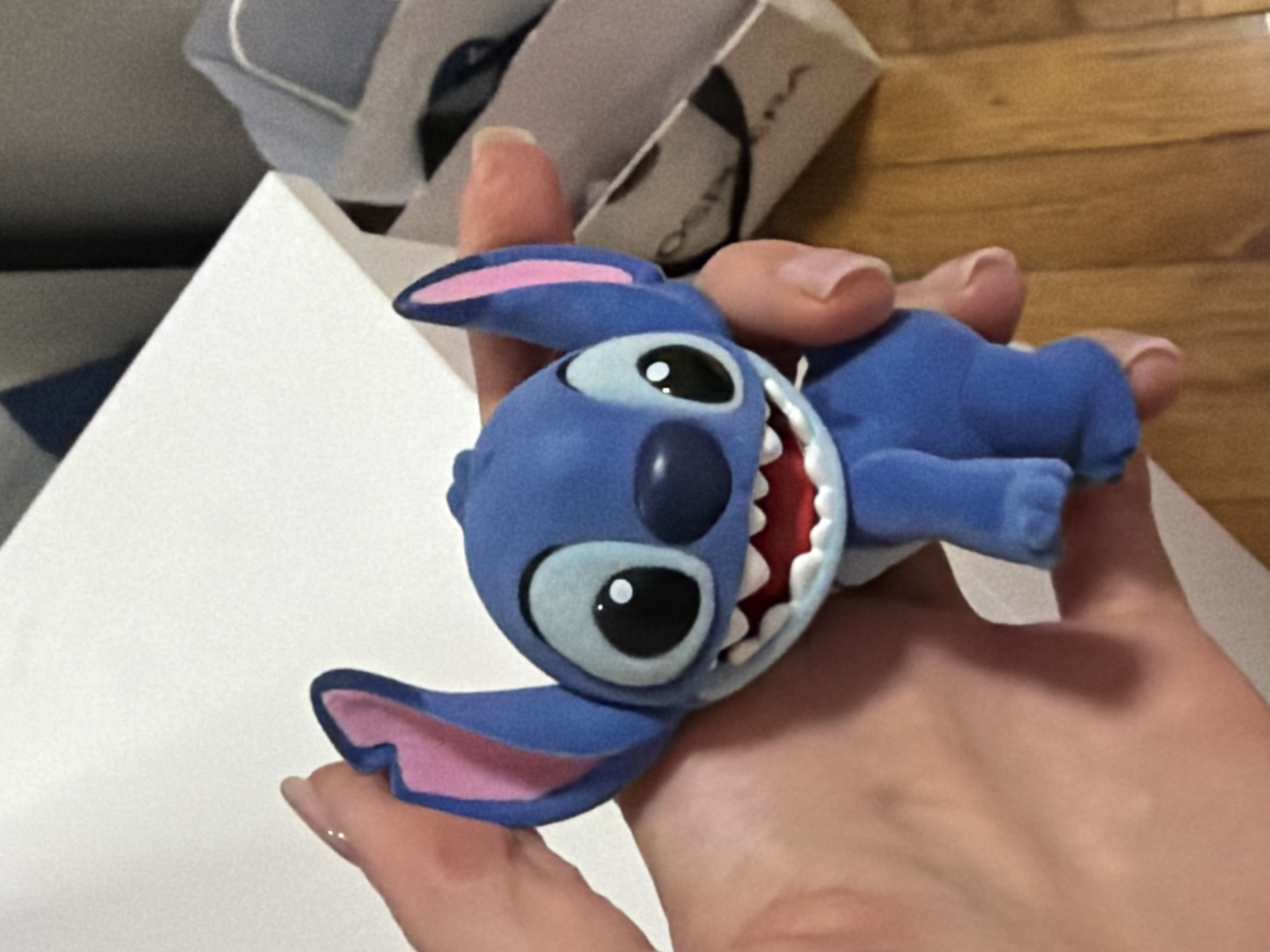 A blue Stitch figurine is held in a hand. The Stitch figurine has large ears, big eyes with black pupils and white highlights, a wide mouth with white teeth and a red tongue. The figurine is positioned slightly to the left and is facing forward. The hand holding the figurine is a fair-skinned hand with manicured nails. In the background, there is a white box, a gray wall, and a wooden floor. To the right of the Stitch figurine is a white paper bag with black writing on it. The scene appears to be indoors. There is no discernible indication of the location being in Moscow, Russia.