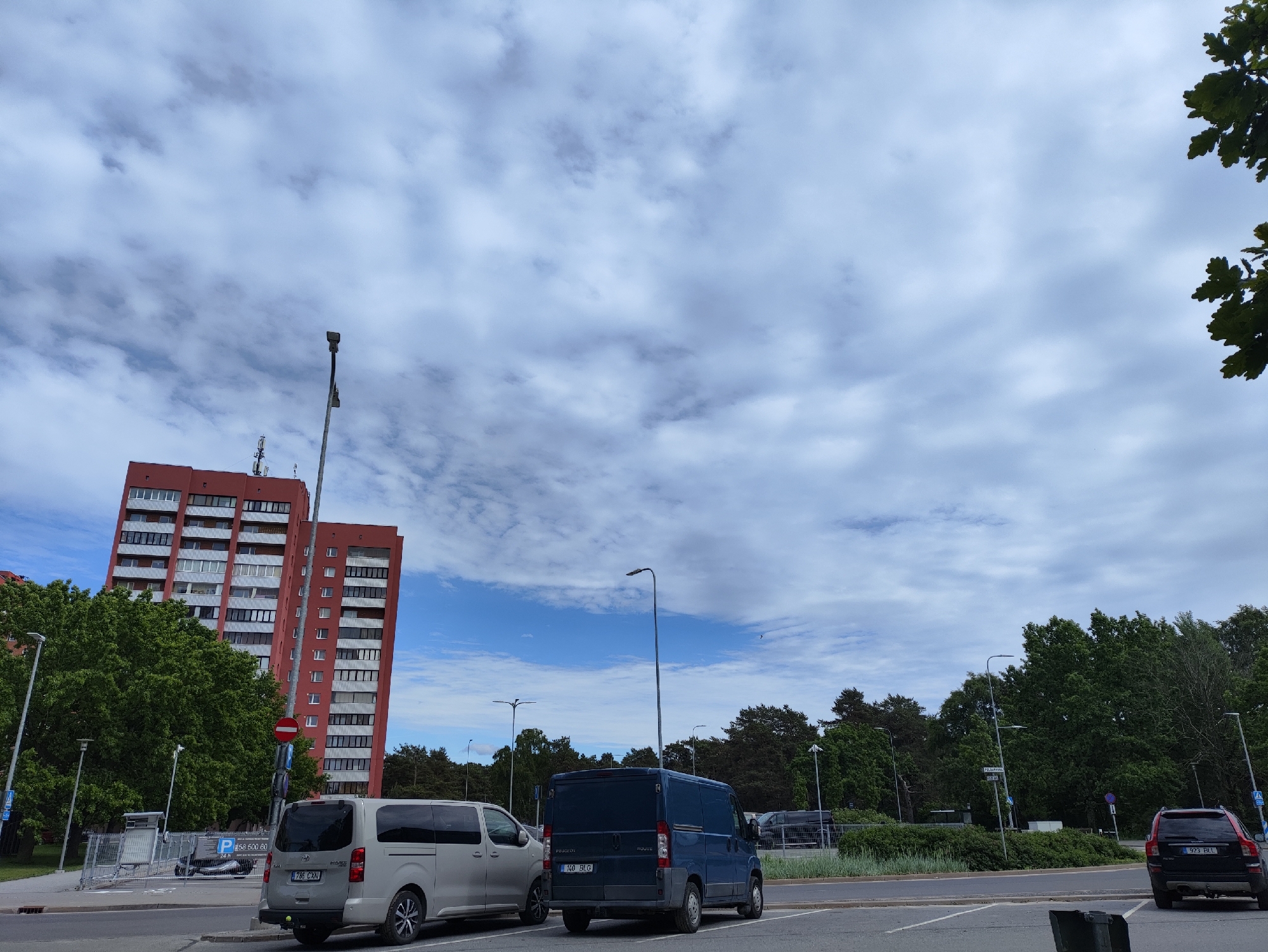 A daytime, eye-level, wide shot depicts a parking lot in Tallinn, Estonia, with a red apartment building and trees in the background. The sky is filled with scattered clouds. In the foreground, there are three parked vehicles: a silver van on the left, a dark blue van in the middle, and a dark red SUV on the right. A street light stands to the right of the silver van, and another one is located in the middle of the image. A traffic light with a red circle and a horizontal bar is visible on the left side of the red building. The red building has many windows and some antennas on its roof. The parking lot is paved with asphalt, and there are white lines marking the parking spaces. The environment appears to be an urban residential area with a mix of buildings and greenery.