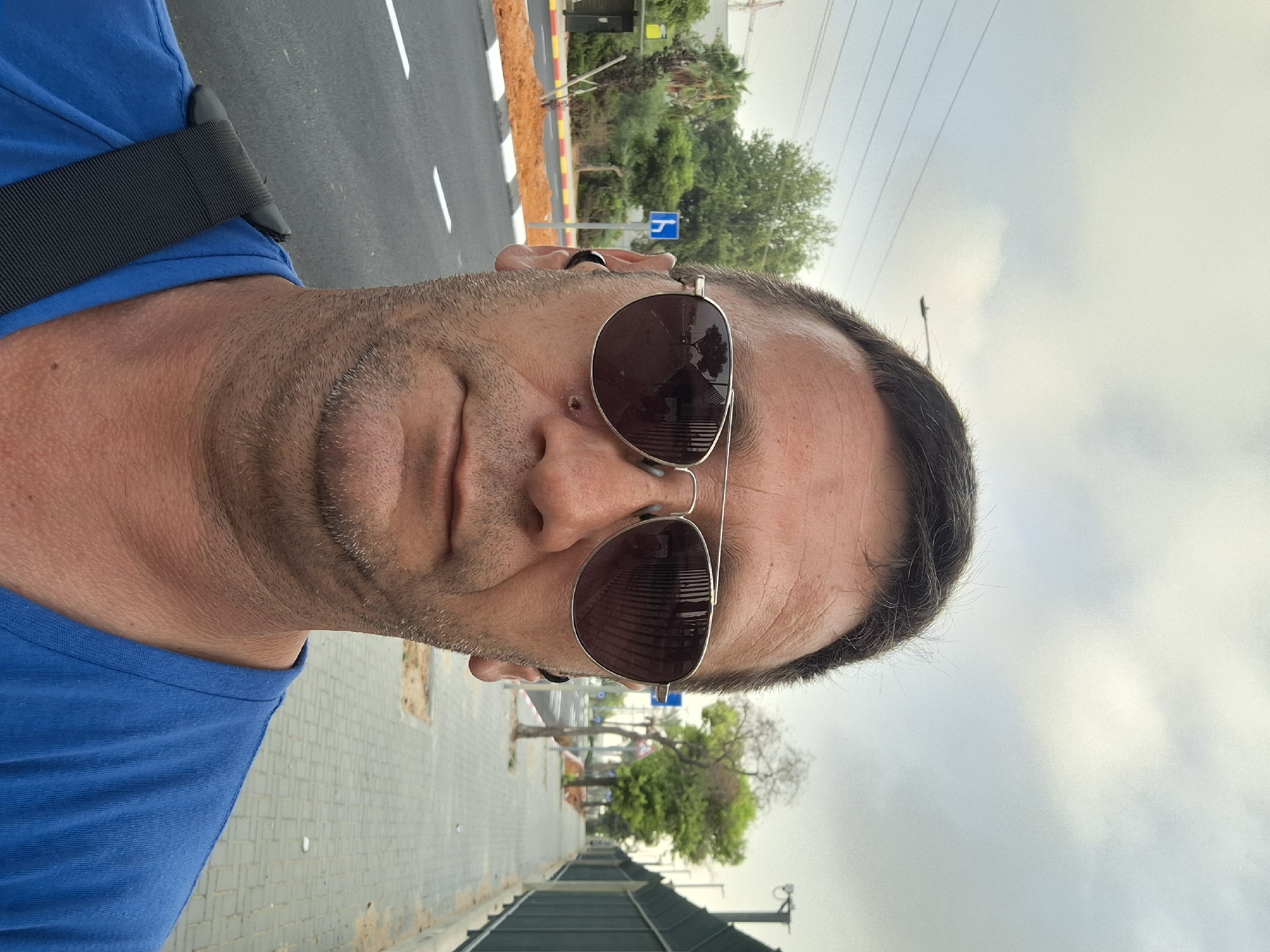 A man wearing aviator sunglasses and a blue t-shirt takes a selfie outdoors. He has short, dark hair and stubble. He is wearing black earbuds. In the background, there is a sidewalk, a street with white lane markings, trees, and a chain-link fence. There is a blue sign with a white icon of a person on a bicycle visible on the side of the road. The sky is overcast. The scene appears to be in Kiryat Gat, Israel, judging by the signage and vegetation.