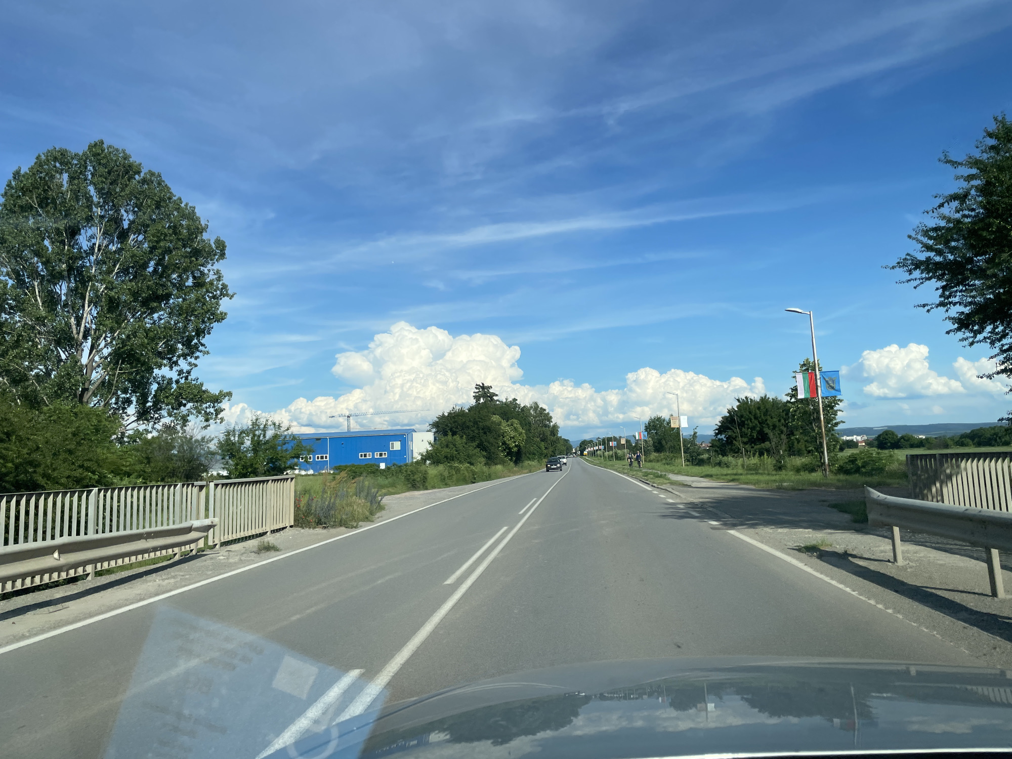 The image shows a road in Musachevo, Bulgaria, on a sunny day with a blue sky and white clouds. The perspective is from inside a vehicle, looking forward. On the left side of the road, there is a metal fence and a large tree. Further down the road on the left, there is a long, blue building with white-framed windows. On the right side of the road, there are more trees, a lamppost with flags, and a few people walking. The road has two lanes in each direction, marked by white lines. There are a few cars on the road. The overall environment appears to be a semi-urban or suburban area.