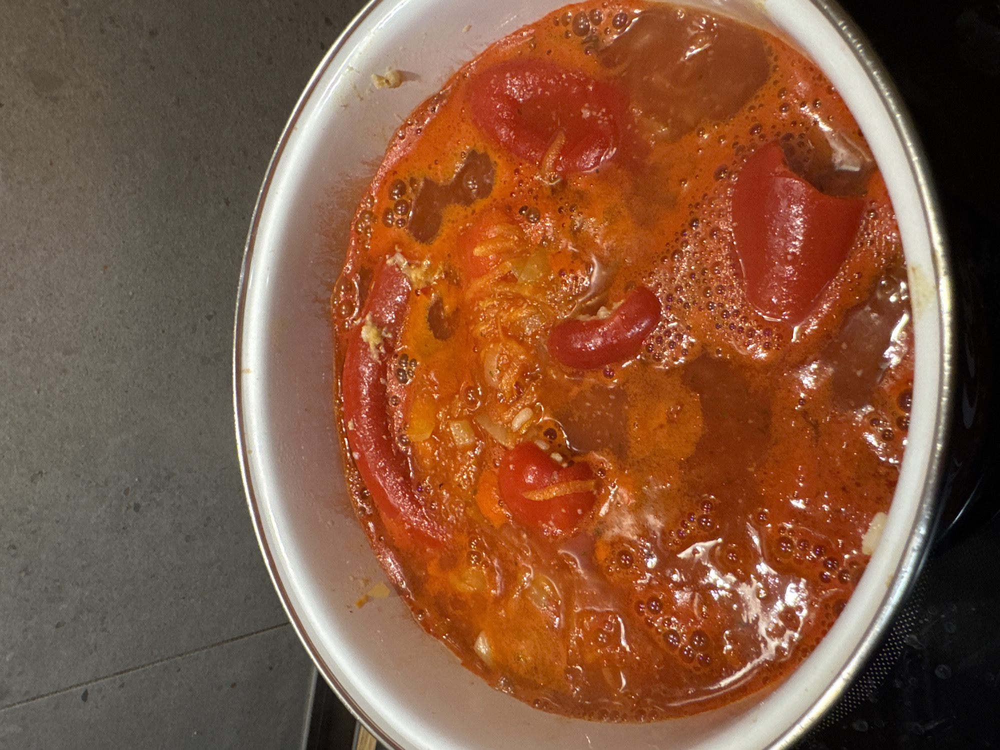 A close-up, overhead shot shows a white enamel pot filled with a bubbling, bright red stew. Large pieces of red pepper and smaller red cherry tomatoes are visible within the liquid, along with bits of what appear to be onions and other vegetables. The stew is actively simmering, with small bubbles breaking the surface in several areas. The pot is resting on a black stovetop. The background features a grey, speckled tiled wall. The location is Unknown, Unknown.