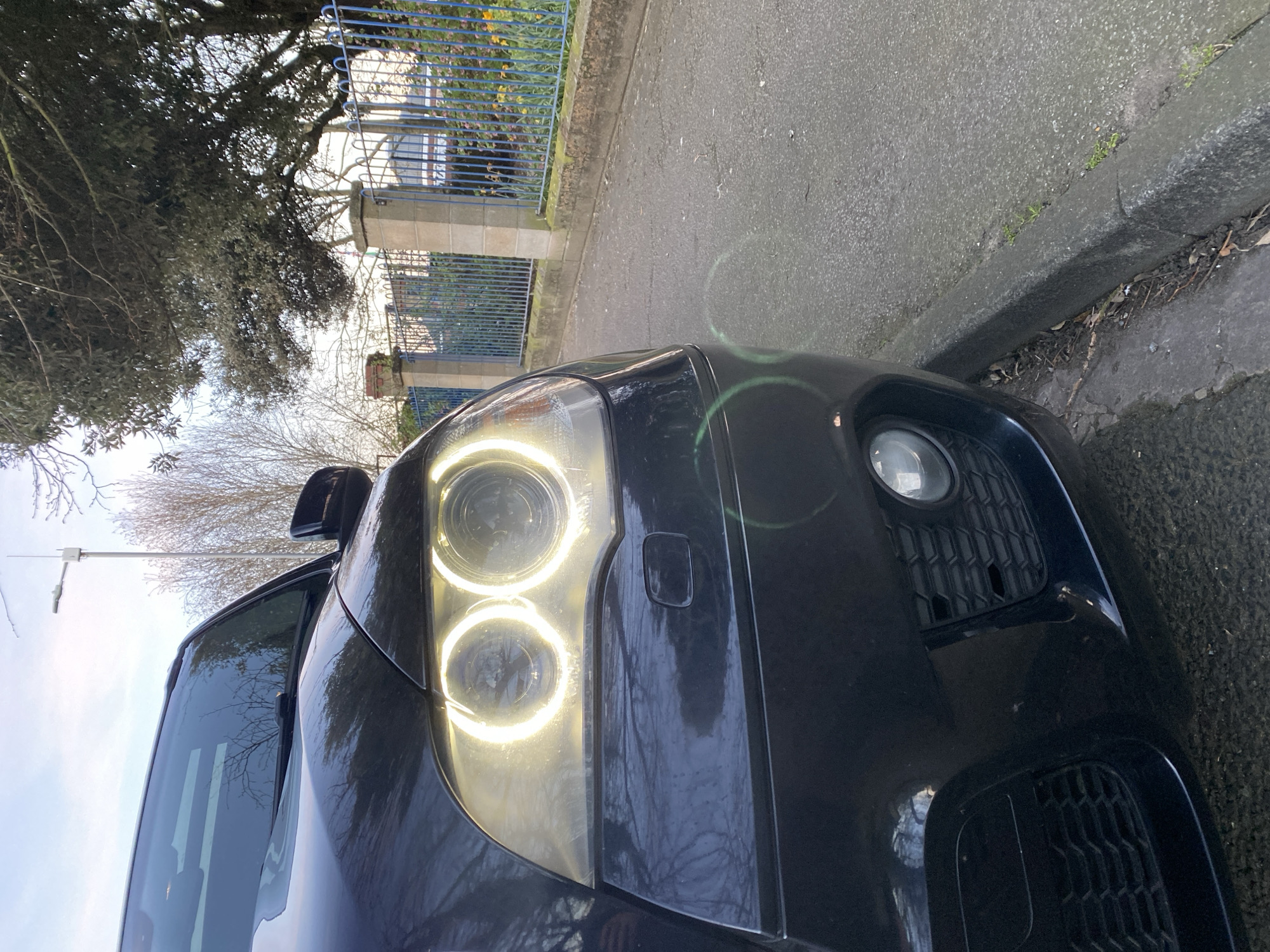 The image depicts the front of a black car parked on a street in East Suffolk, United Kingdom. The car's headlights are illuminated, showing a double-ring design. The street is paved and has a curb along the right edge. A tree and a blue metal fence are visible in the background. There are no people visible in the image.
