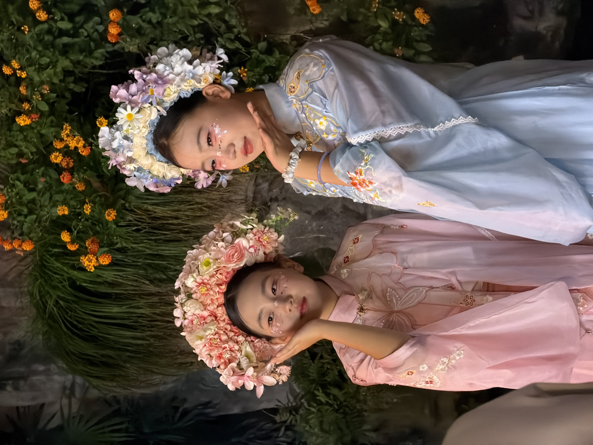The image shows two young girls in traditional Chinese attire, posing in front of a lush green backdrop with orange flowers. The girl on the left wears a pink hanfu with intricate embroidery and a large floral headdress. She has her hand resting on her cheek. The girl on the right wears a light blue hanfu, also adorned with embroidery and a floral headdress. She is blowing a kiss. Both girls have light makeup with glitter on their faces. The setting appears to be outdoors, possibly in a garden or park. The specific location in Yuzhong District, China, is not determinable from the image alone.