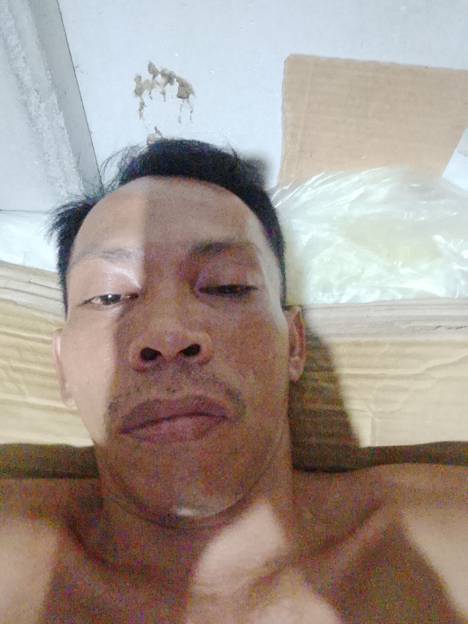 The image shows a close-up of a shirtless man's face. He has short, dark hair and appears to be lying down. One of his eyes is open, while the other is partially closed. The lighting is uneven, casting a shadow across half of his face. In the background, there is a white ceiling with some staining, a cardboard box, and a plastic bag filled with white material, possibly packaging. The background also shows a light yellow surface. Based on the prompt, the location is Bukit Ketok, Indonesia, but no elements in the image explicitly confirm the location.