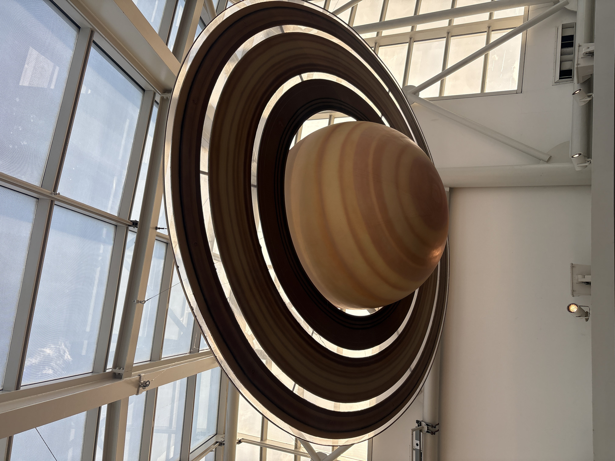 This image depicts a model of the planet Saturn with its rings, suspended indoors, possibly in a museum or exhibit. The planet is striped in shades of beige and brown, and its rings are composed of concentric dark brown circles. To the left, a wall with multiple windows and white metal framework is visible. To the right, a plain white wall is seen. The scene is well-lit with natural light streaming through the windows. It is likely located in the city of Chicago, United States of America.