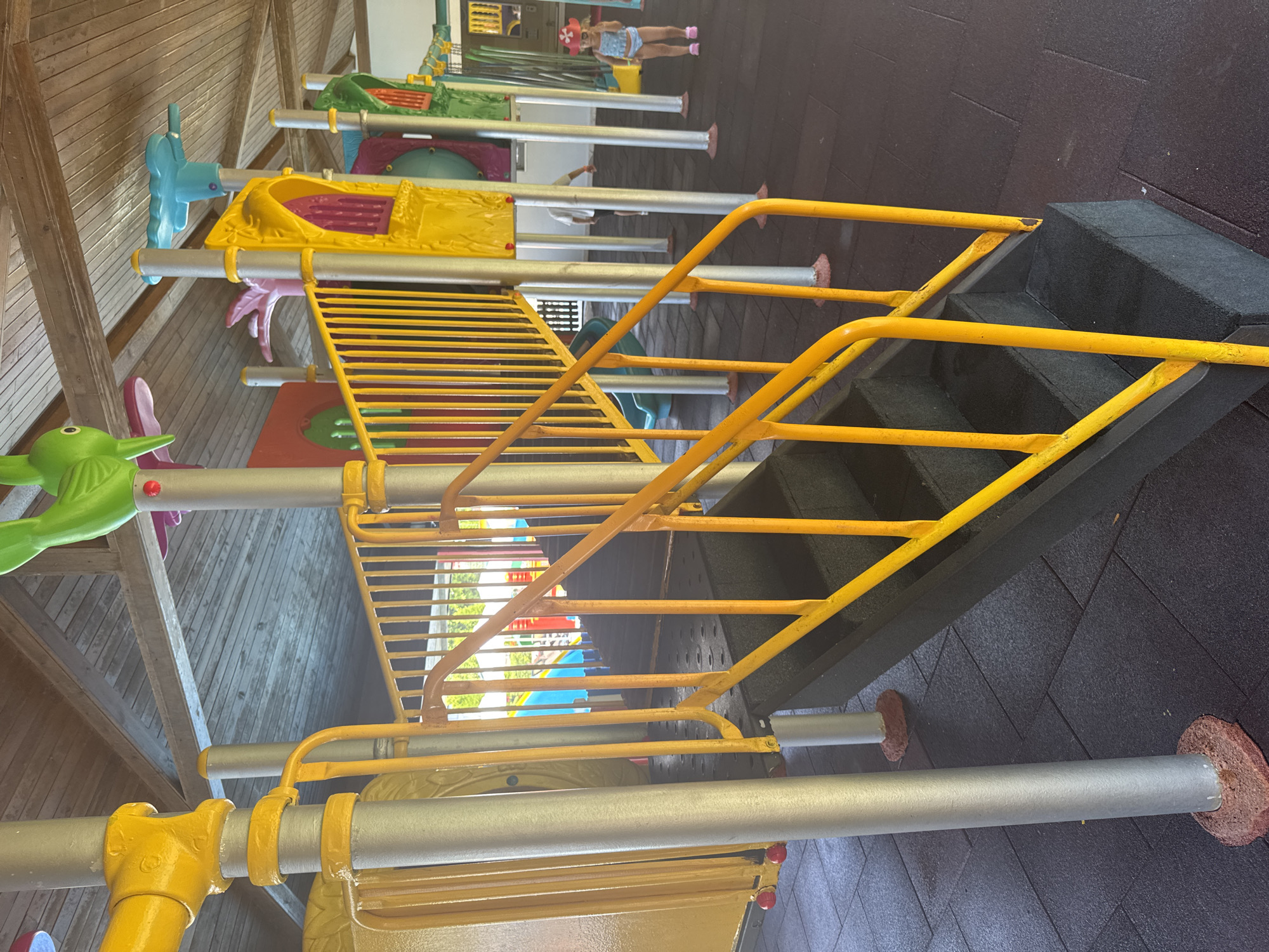 The image shows an outdoor children's playground in Aksu, Turkey. The playground features a yellow and gray metal structure with stairs and railings, along with colorful plastic elements like slides and decorative figures. The ground is covered with dark, rubberized safety tiles. A young child wearing a blue swimsuit and a red-and-white hat is visible on the right side of the image, standing on the rubberized tiles. Another person, partially visible, is walking in the background. The playground appears to be partially covered by a wooden roof.
