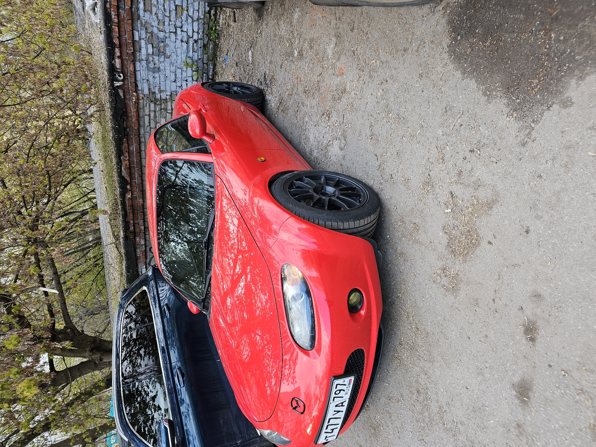 The image captures a static outdoor scene featuring two parked cars next to a brick wall and trees, likely in Khimki, Russia.
The primary subject is a vibrant red Mazda Miata (MX-5), appearing to be an NC generation model with a hardtop. It is parked on an unpaved, somewhat dirty ground surface that transitions to a paved area further back. The car is equipped with distinctive black multi-spoke aftermarket wheels. A yellow-tinted fog light is visible on the right side of its front bumper, and the Mazda emblem is centered on the hood. The car's front license plate is clearly visible, displaying "T477YA797 RUS," which identifies it as registered in Russia, with "797" being a Moscow region code. The reflections on its glossy red paint suggest bright, sunny daylight conditions.
To the left of the red Mazda, a dark blue or black car is partially visible, parked parallel. Only its side, windows, and part of its roof are seen. The reflection of the trees is noticeable on its windows.
In the background, behind the cars, there is a weathered brick wall, parts of which appear to have peeling paint or plaster. Above the wall, a line of deciduous trees with fresh green leaves indicates that the photo was taken during spring or early summer. The ground is a mix of asphalt and dirt, scattered with small debris and dried leaves.
No people are visible, and no specific activity is taking place other than the cars being parked. The overall impression is that of a quiet street or parking area during the daytime.