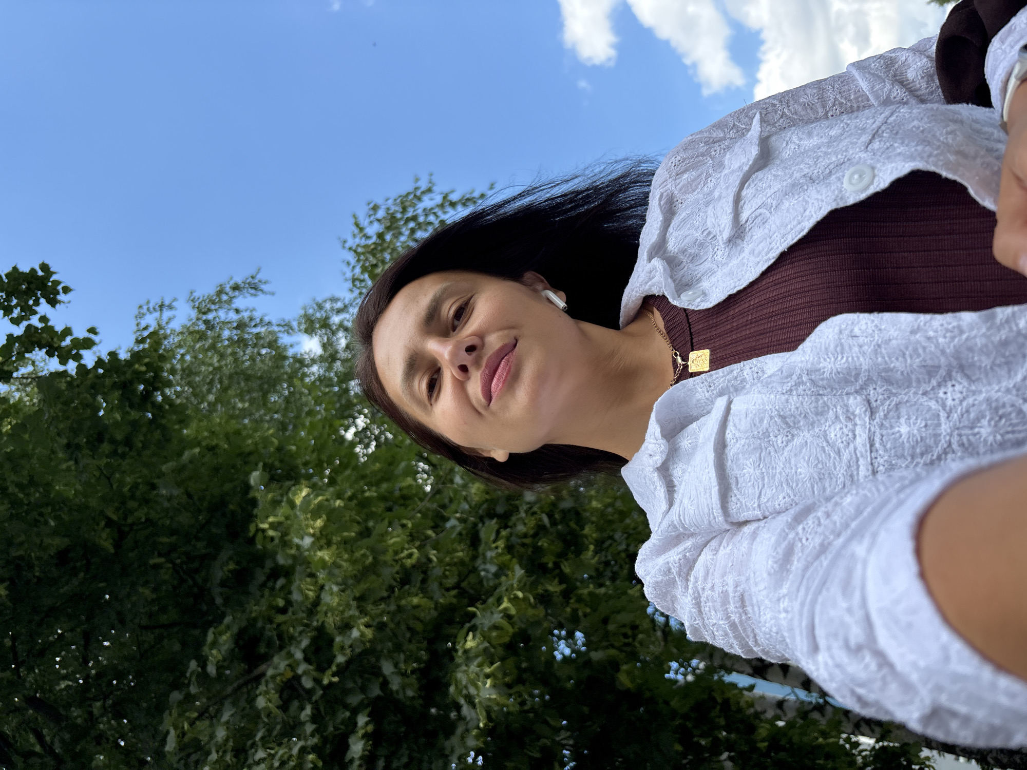 The image is a low-angle selfie of a woman outdoors, with lush green trees and a blue sky with scattered white clouds in the background. She is looking upwards towards the camera, with a slight smile on her face. She has long, dark hair and is wearing a white, textured blouse over a dark brown top. A gold necklace with a square pendant is visible. She is also wearing white wireless earbuds. The lighting suggests it is daytime, likely sunny, as indicated by the bright sky and the illumination on her face. The overall mood appears casual and relaxed. No visible text is present in the image. The provided location context is Chaikovsky, Russia.