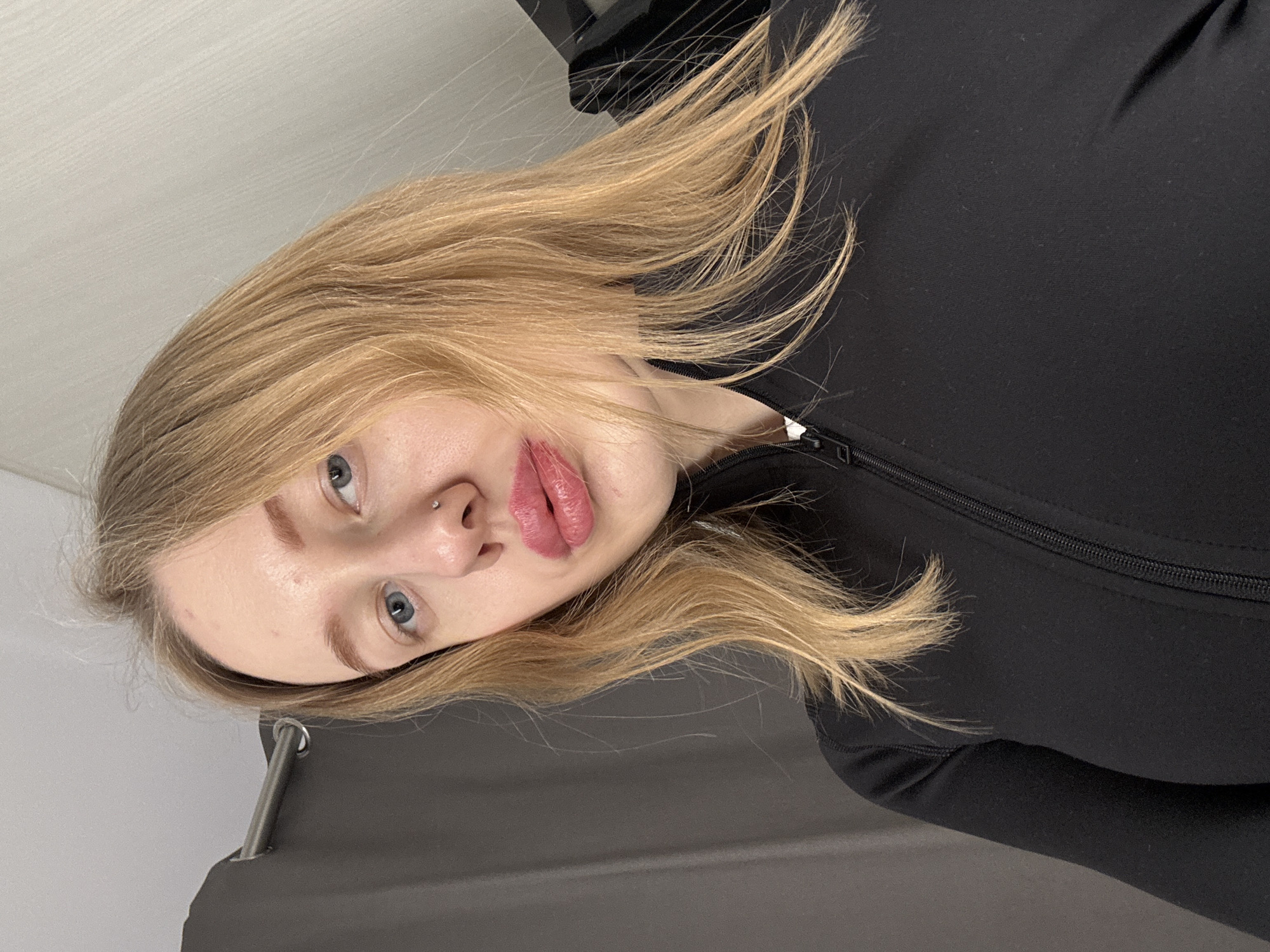 A close-up, front-facing portrait depicts a female subject with blonde hair, blue eyes, and a light complexion. The subject presents with a small, reflective stud piercing in her left nostril and wears glossy, pink-red lipstick. She is dressed in a black, long-sleeved garment featuring a zippered front closure. The visible background indicates an indoor setting, composed of a dark gray fabric curtain suspended from a silver-colored rod on the subject's left, and a light gray textured wall surface on the subject's right. The ceiling, light in color, is visible at the top of the frame. No specific environmental markers are present within the image to identify the location as Rostov-on-Don, Russia.