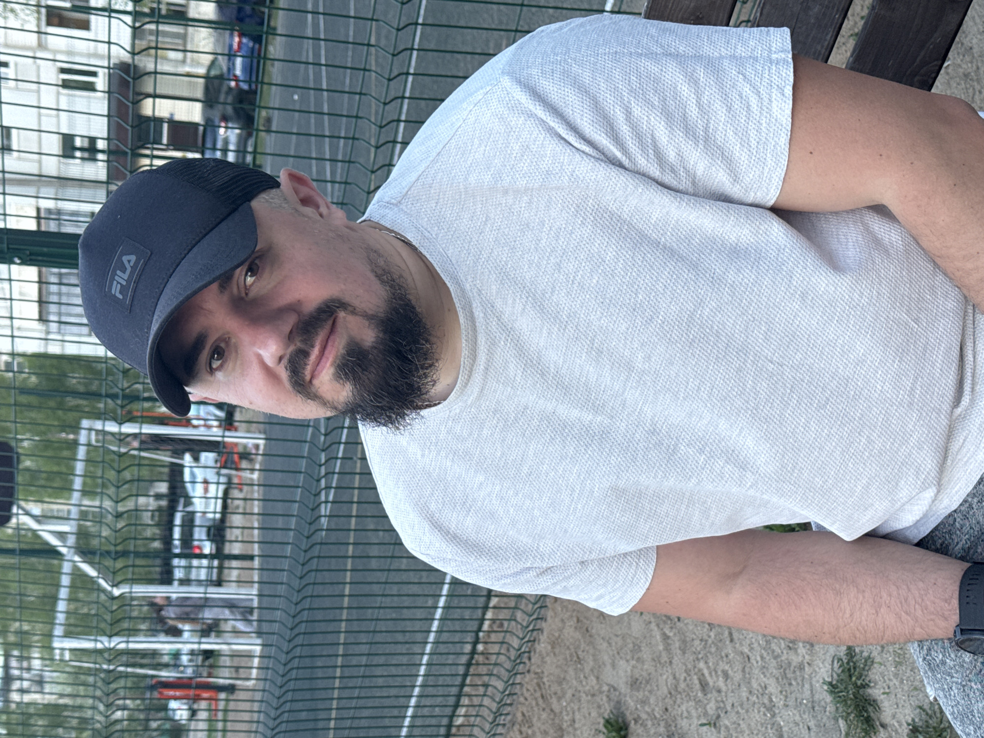 A medium shot depicts a man sitting outdoors. He is wearing a white t-shirt and a dark-colored Fila baseball cap. The man has a dark beard and is looking towards the camera. A fence is visible in the background, behind which is a building. It is daytime. The image was taken in Chaikovsky, Russia.