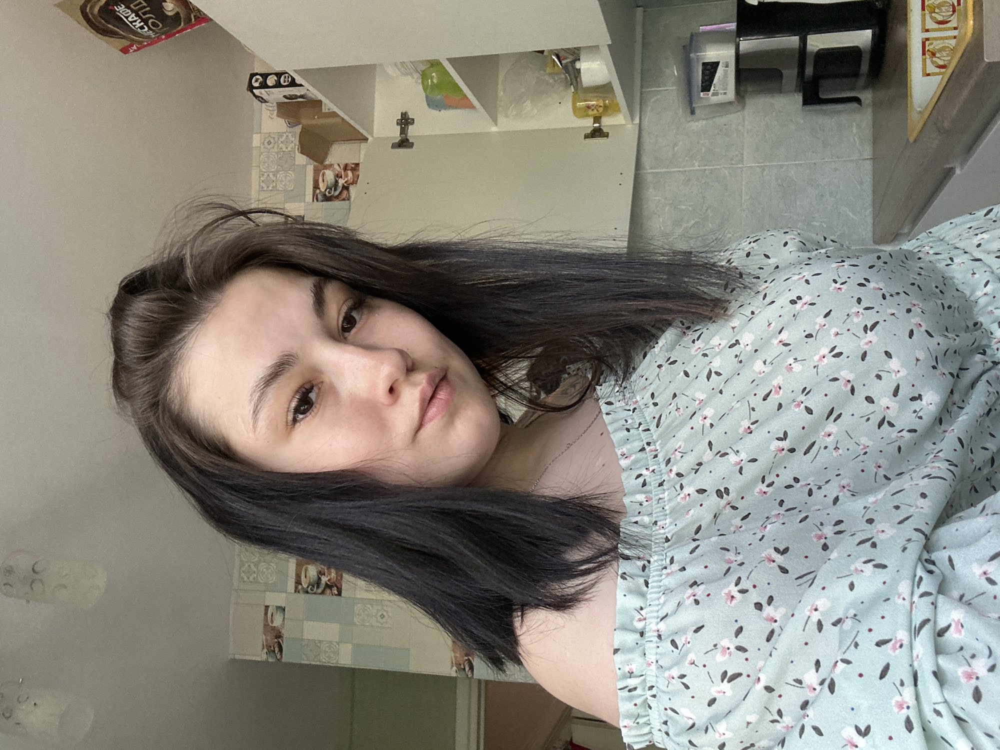A woman is in a kitchen with light blue flowered shirt. The kitchen has white cabinets. The woman is looking at the camera. The kitchen walls have blue and white tiles. There is a coffee box on the top left. The location is in Chaikovsky, Russia.