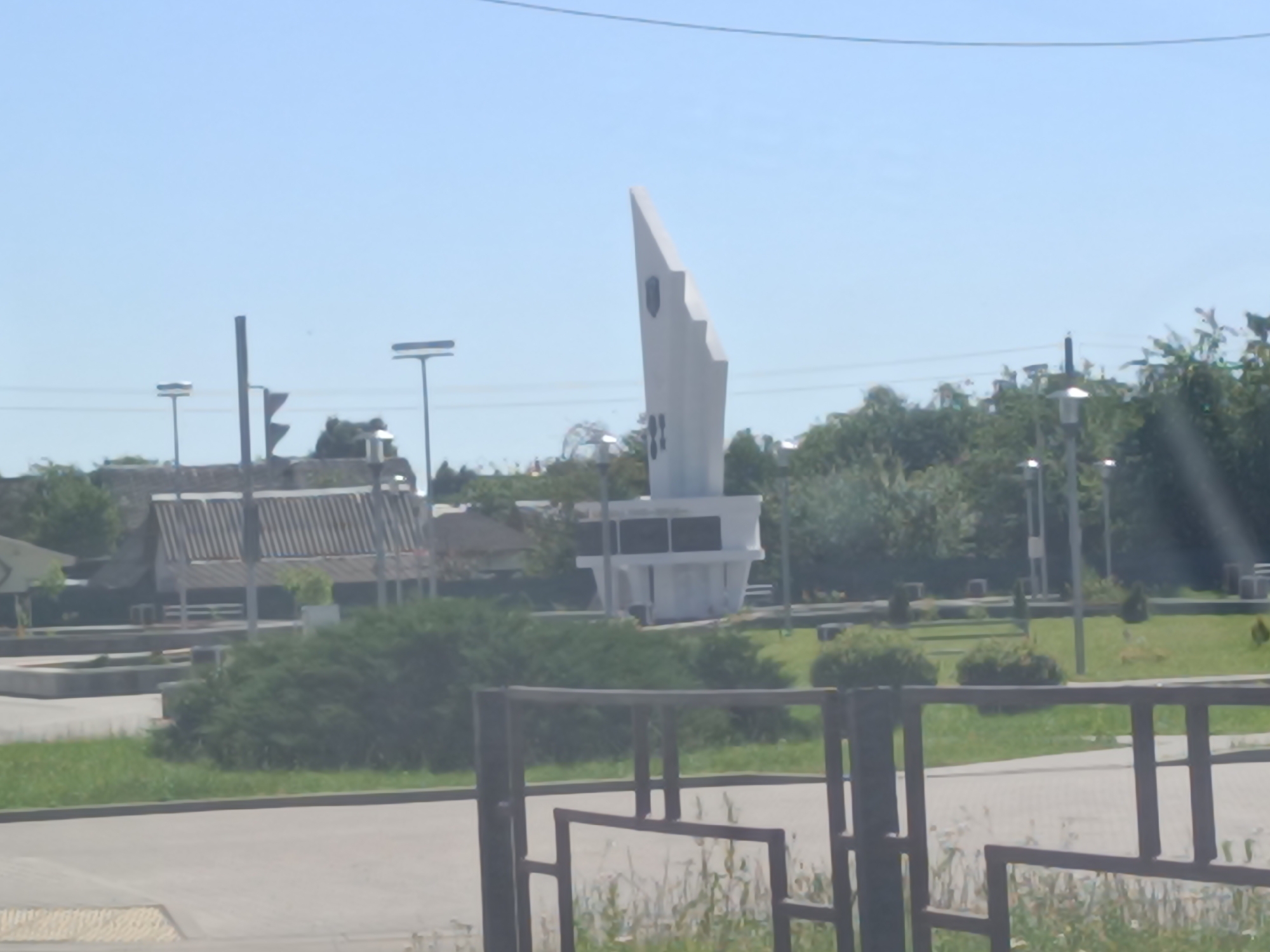 The image depicts a monument in the city of Orsha, Belarus. The monument is a tall, white, abstract sculpture with sharp angles, resembling a stylized flame or shard. It stands in a park-like setting with green grass and manicured bushes. In the background, there is a covered structure with a striped roof, possibly a pavilion or seating area, and several lamp posts. The sky is clear and blue, indicating a daytime scene. There are no people or animals visible in the image. The foreground shows a paved area and a dark metal fence. The overall impression is of a public monument in a well-maintained outdoor space.