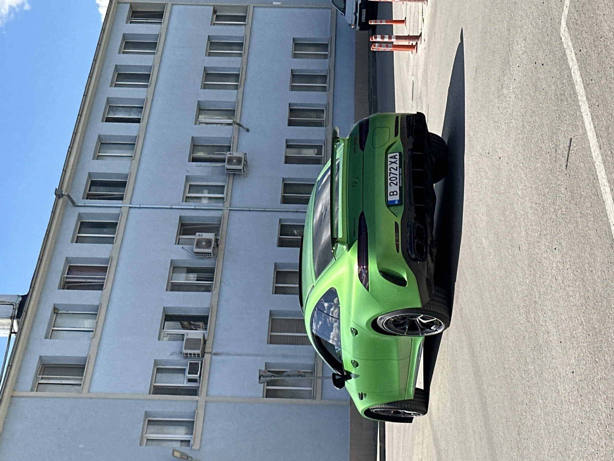 A green sports car is parked on the side of a building. The car has a Bulgarian license plate that reads "B 2072 XA." The building is a light blue color and has many windows and air conditioning units. A few orange traffic cones are also visible. The location appears to be an urban environment in Bulgaria. Due to the license plate beginning with B, it is likely that this photo was taken in or near Sofia, Bulgaria rather than Varna.