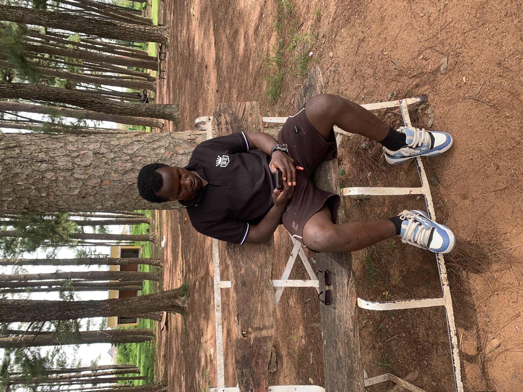 The image depicts a Black man sitting on a wooden bench in a park-like setting with tall trees. The man is wearing a black polo shirt with white trim and dark shorts, and is looking down at his mobile phone, which he is holding in his hands. He is also wearing blue and white sneakers and black socks. A pair of sunglasses rests on the bench to his left. The ground is covered in reddish-brown dirt and fallen pine needles. In the background, there is a small, yellow building with dark windows and a line of trees. The environment suggests a location in Jos, Nigeria, a city known for its pine forests and cooler climate.