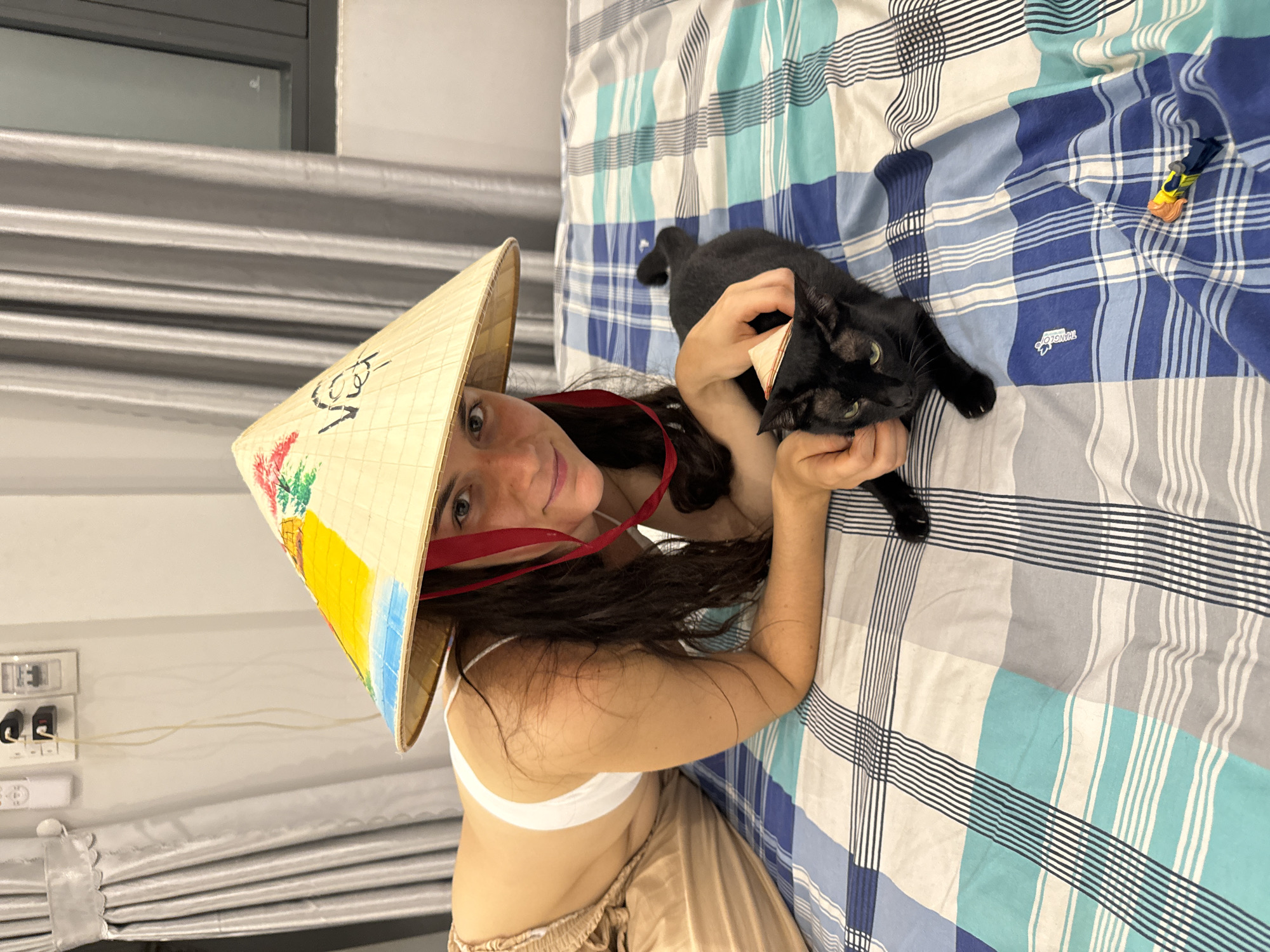 A woman wearing a Vietnamese conical hat poses with a black cat on a bed in a room in Mũi Né, Vietnam. The woman lies on the bed, supporting herself with her arms. She wears a white top and beige bottoms. The conical hat is decorated with colorful painted designs. The black cat lies between her hands. The bed has a blue and white checkered pattern bedsheet with a toy car in the upper right corner. A window and electrical outlets are visible on the wall behind the woman.