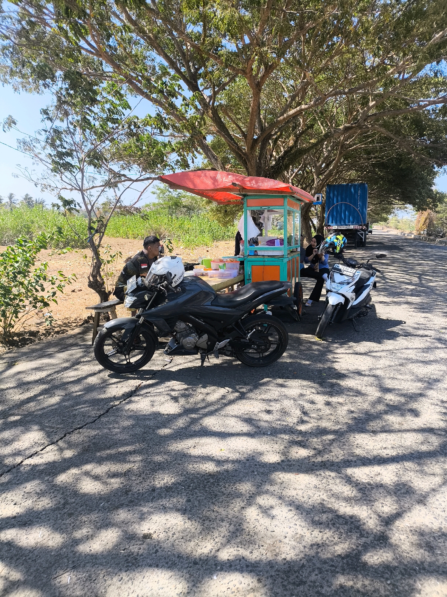 A roadside scene in Kwandang, Indonesia, features a mobile food cart with a red canopy set up beneath a large tree. Two motorcycles are parked nearby. A man in a black shirt with white accents sits on a wooden stool to the left of the food cart, facing away from the camera. A white helmet rests on the handlebars of a black motorcycle parked directly in front of the cart. Another motorcycle, white with black accents, is parked to the right of the cart. Two women are visible interacting with the food cart. One woman, wearing a dark hijab and black clothing, sits on the edge of the cart's counter. Another woman, partially obscured, stands behind the counter. In the background, a blue truck is parked on the right side of the road, and vegetation lines the left side of the road. The ground is paved and shows dappled sunlight and shadows from the trees.