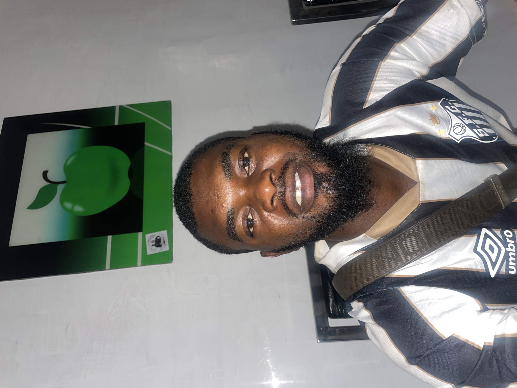 A man with a beard is in the frame, wearing a black and white striped soccer jersey, possibly in a domestic setting. A green apple graphic in a black frame hangs on the wall to his left. The man has a dark complexion and is smiling slightly. A brown strap is visible across his chest. The location is not explicitly specified, however, the prompt implies a location within the city of Jos, Nigeria.